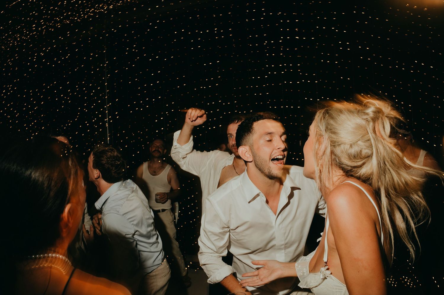 Groom celebrating joyfully on the dance floor at a lively wedding reception under fairy lights.