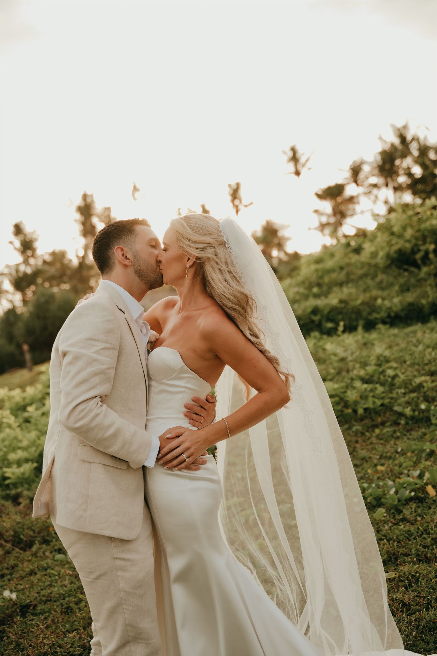 Bride and groom share a romantic sunset kiss, with her veil flowing in a lush tropical garden.