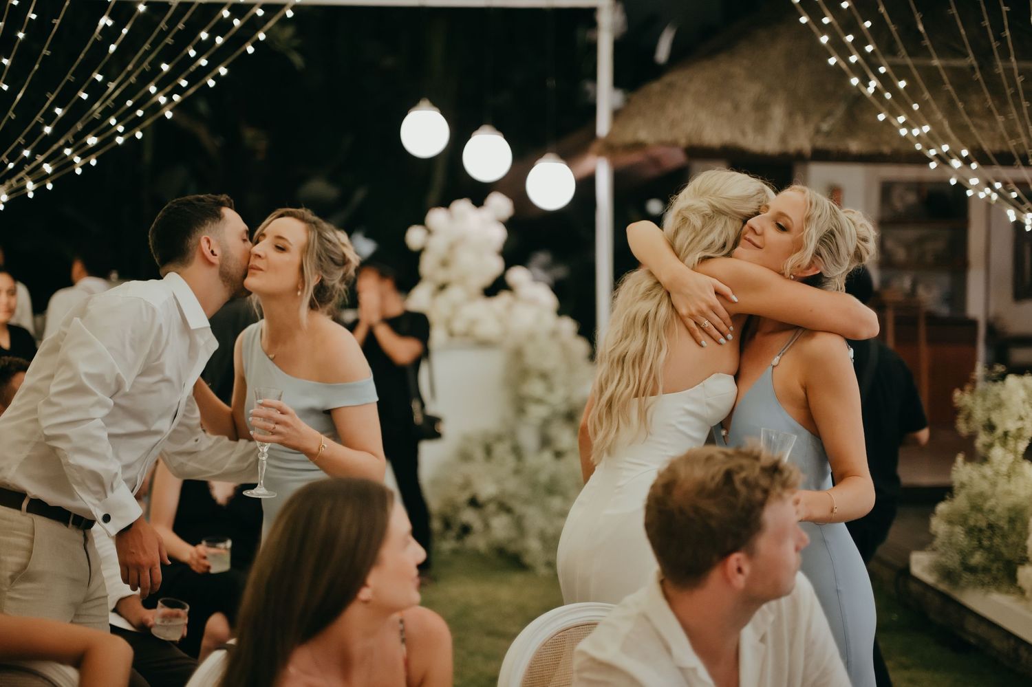 Wedding guests embracing and celebrating at an outdoor reception with string lights and floral backdrop.