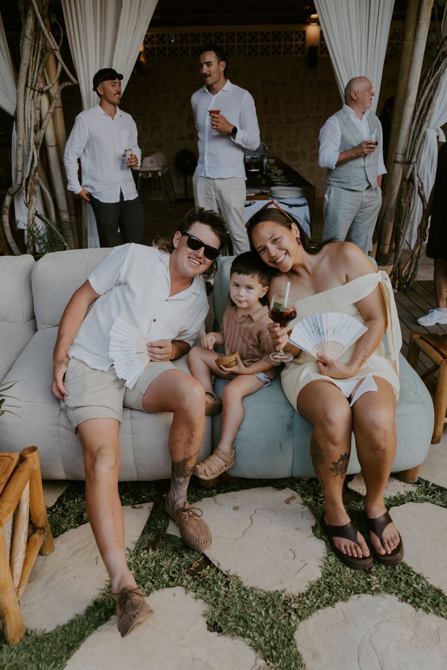 Happy family with young child relaxes on outdoor sofa during tropical wedding reception celebration.