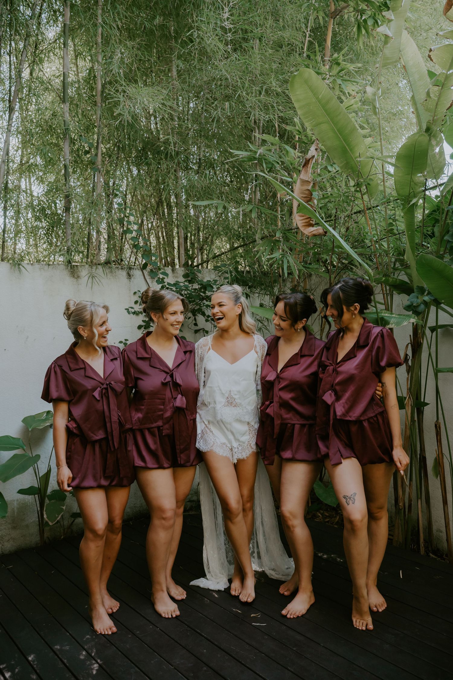 Bride and bridesmaids in burgundy satin rompers laughing together on a wooden deck surrounded by lush tropical greenery.