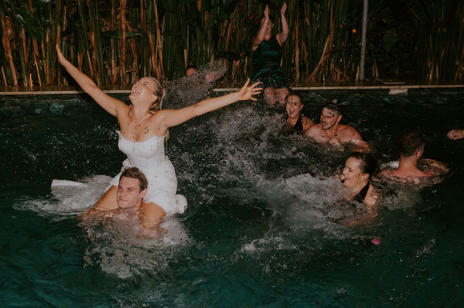 Bride in wedding dress celebrates with guests splashing in a pool during festive wedding after-party.