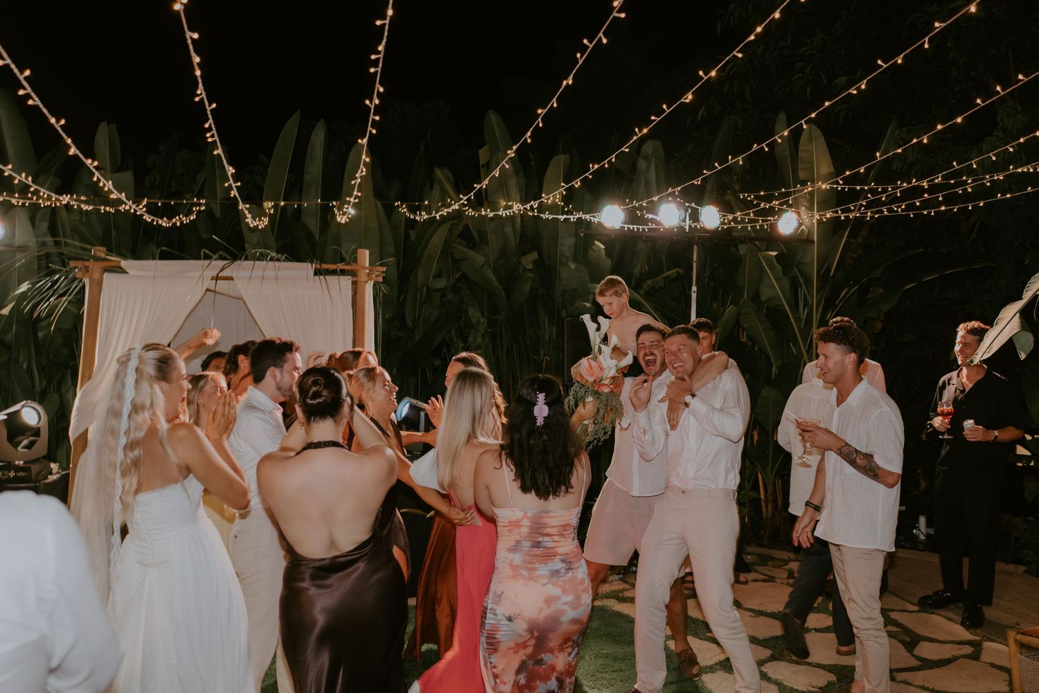Joyful wedding guests dancing and celebrating with the newlyweds under twinkling string lights at a tropical venue.