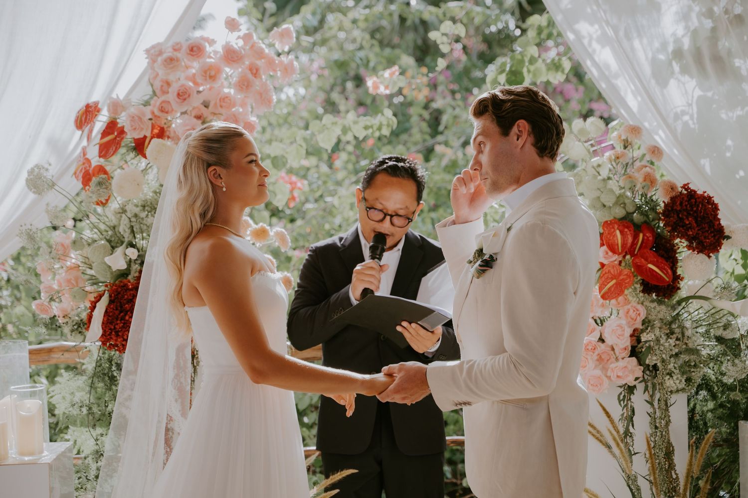 Emotional groom wipes tear during outdoor wedding ceremony with bride and officiant amid floral backdrop.