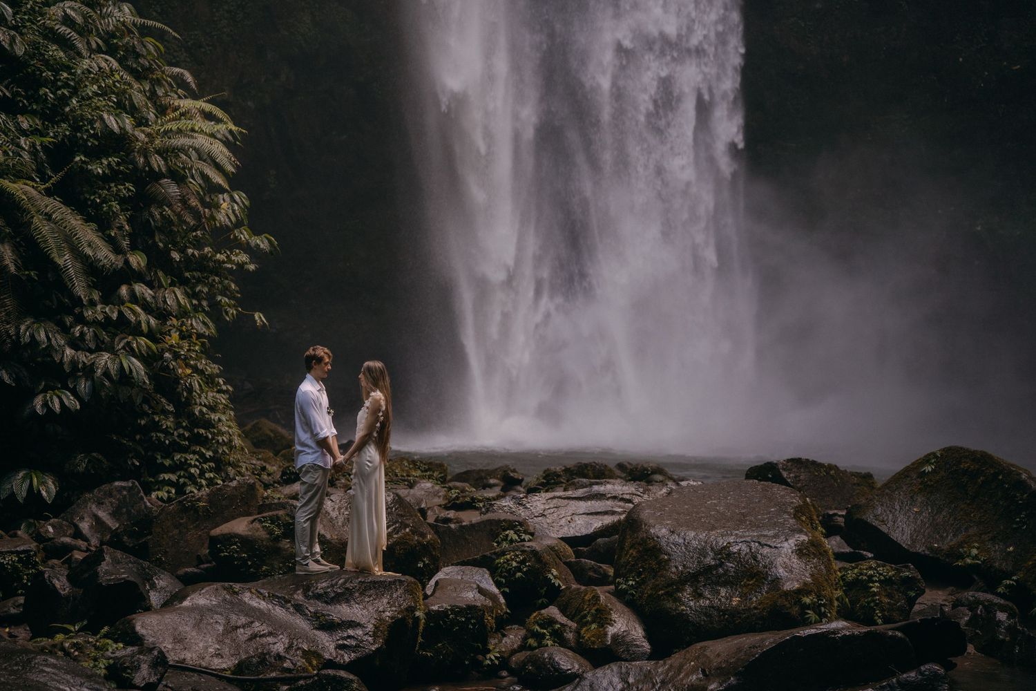 A couple stands on rocky terrain near a majestic waterfall in a moody, atmospheric forest landscape.