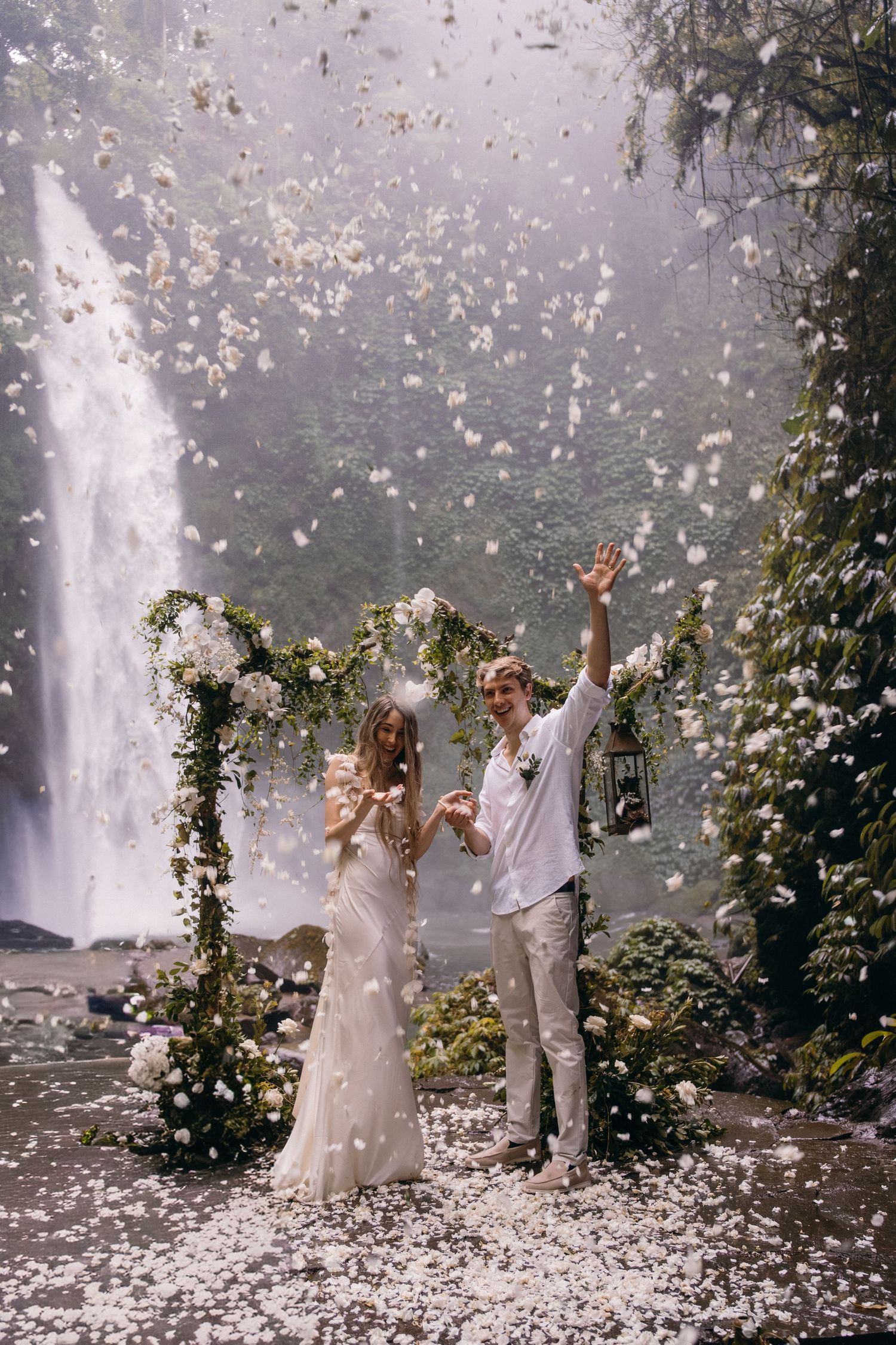 A couple celebrates their wedding ceremony under a floral arch near a waterfall as white petals rain down around them.