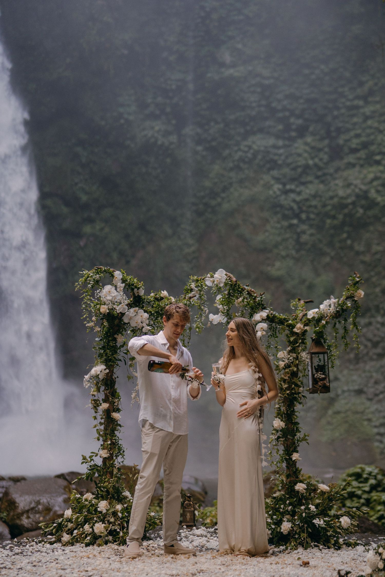 A couple shares a celebratory toast under a floral arch with a misty waterfall backdrop in Hawaii.