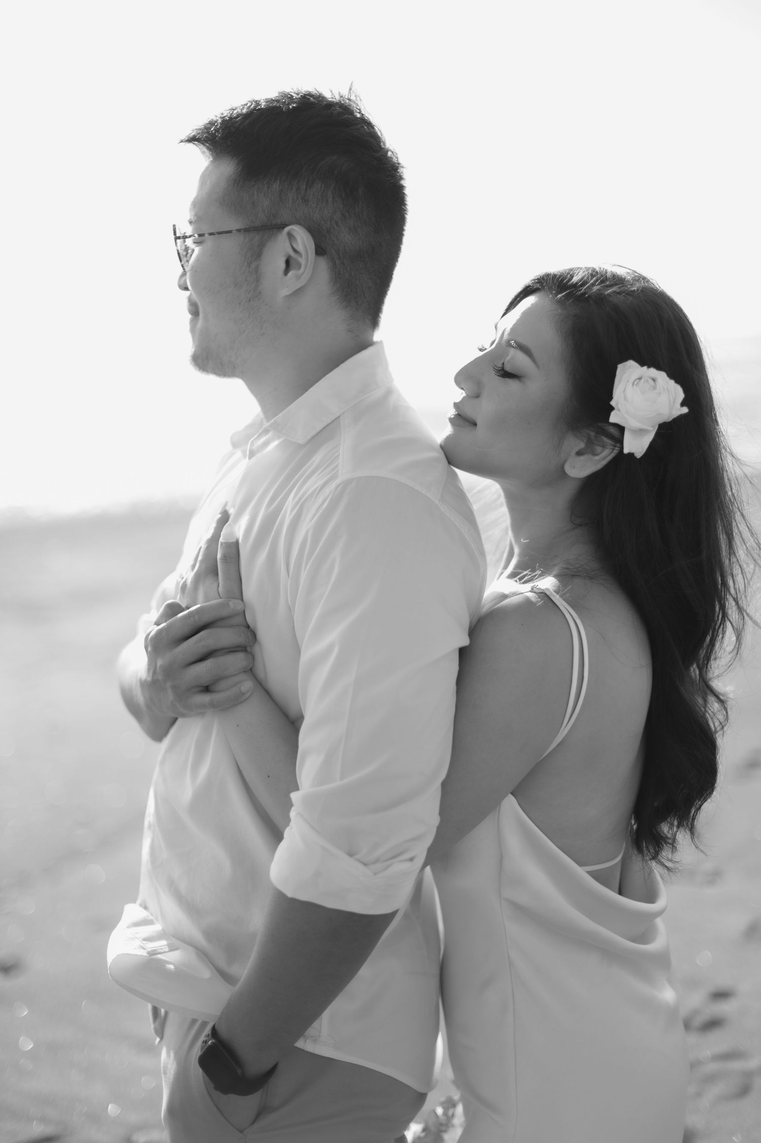 Black and white photo of a couple embracing on a beach, woman with flower in hair resting her head on mans back.