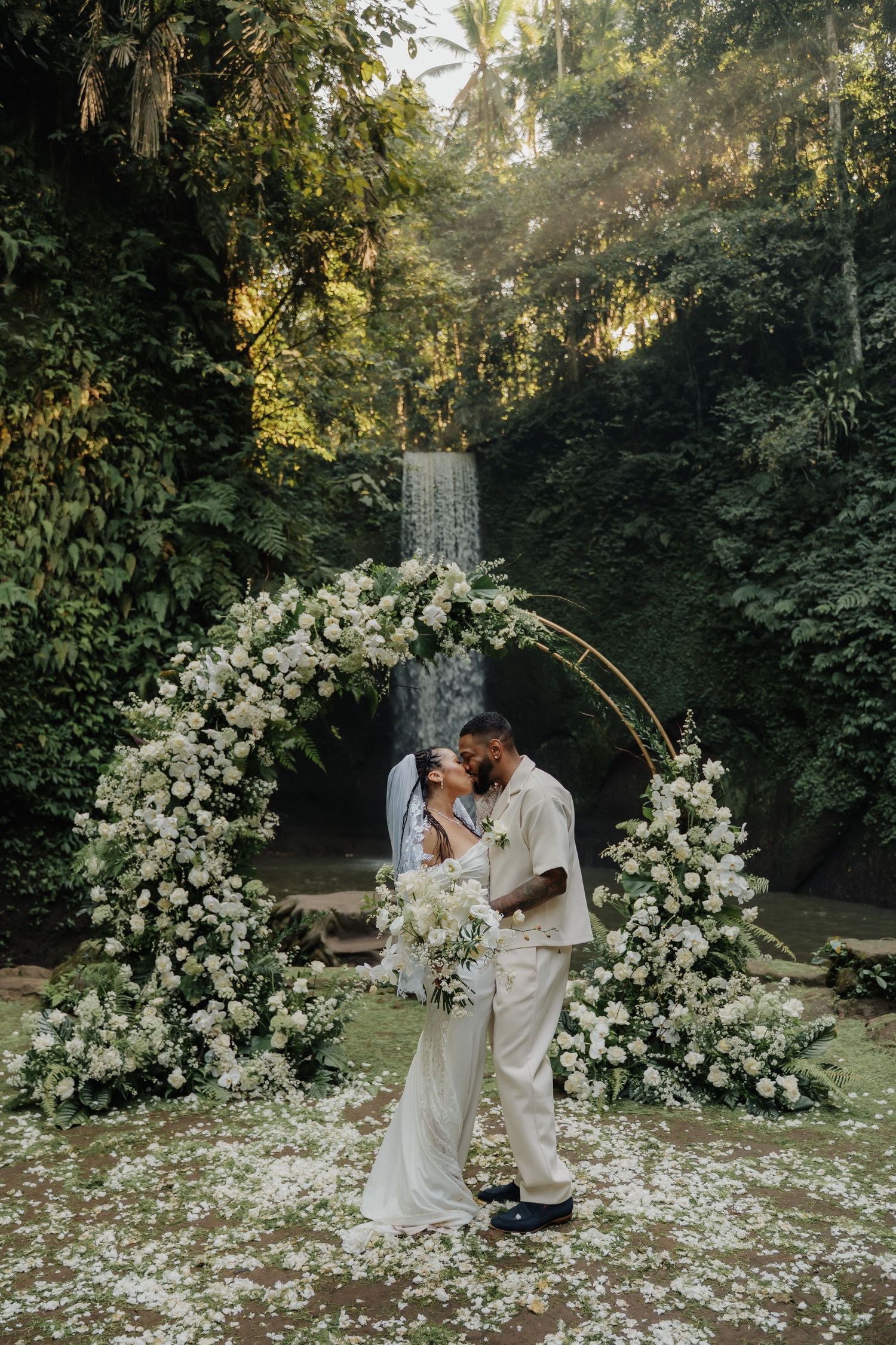 Newlyweds sharing first kiss under white circular floral arch with Bali waterfall backdrop.