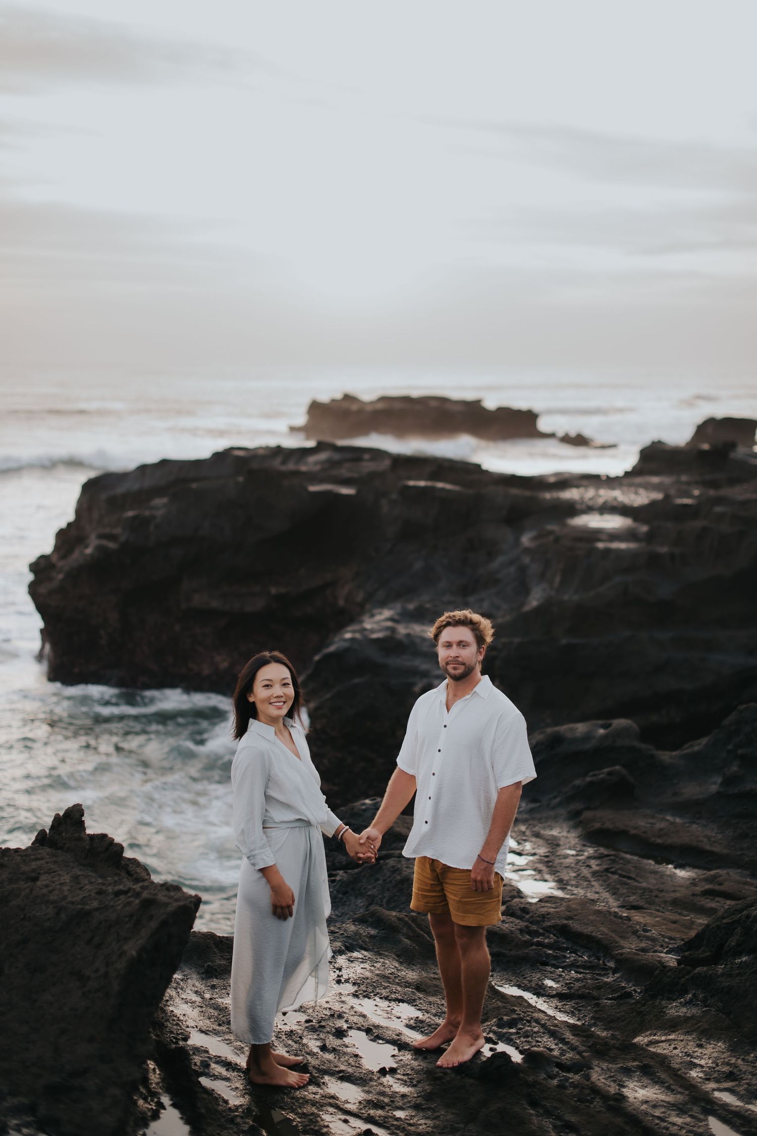 A couple stands together holding hands on dramatic rocky coastal cliffs at sunset with ocean waves crashing below.