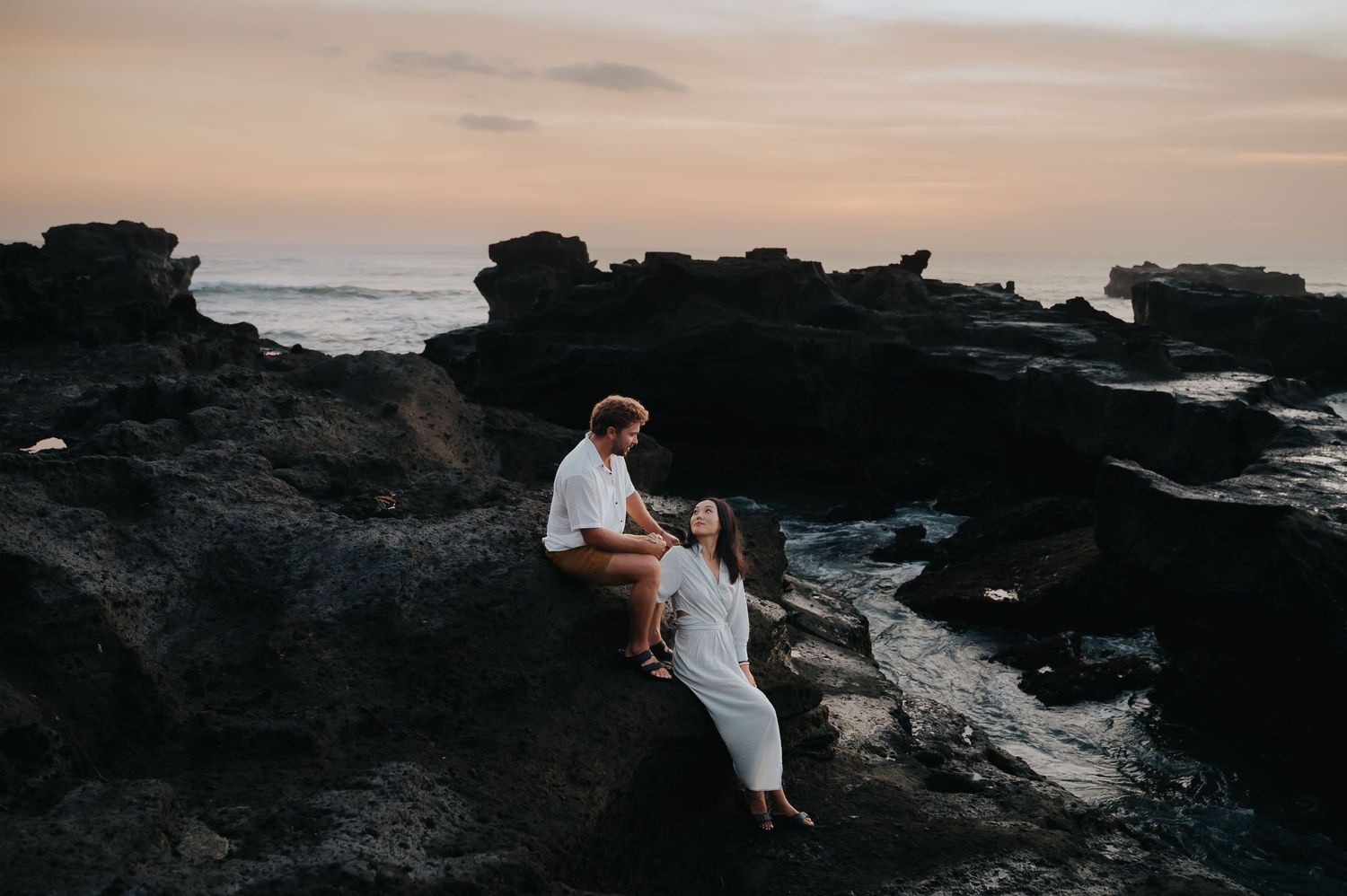 Romantic couple shares intimate moments during sunset photoshoot on dramatic black lava rocks by the ocean.