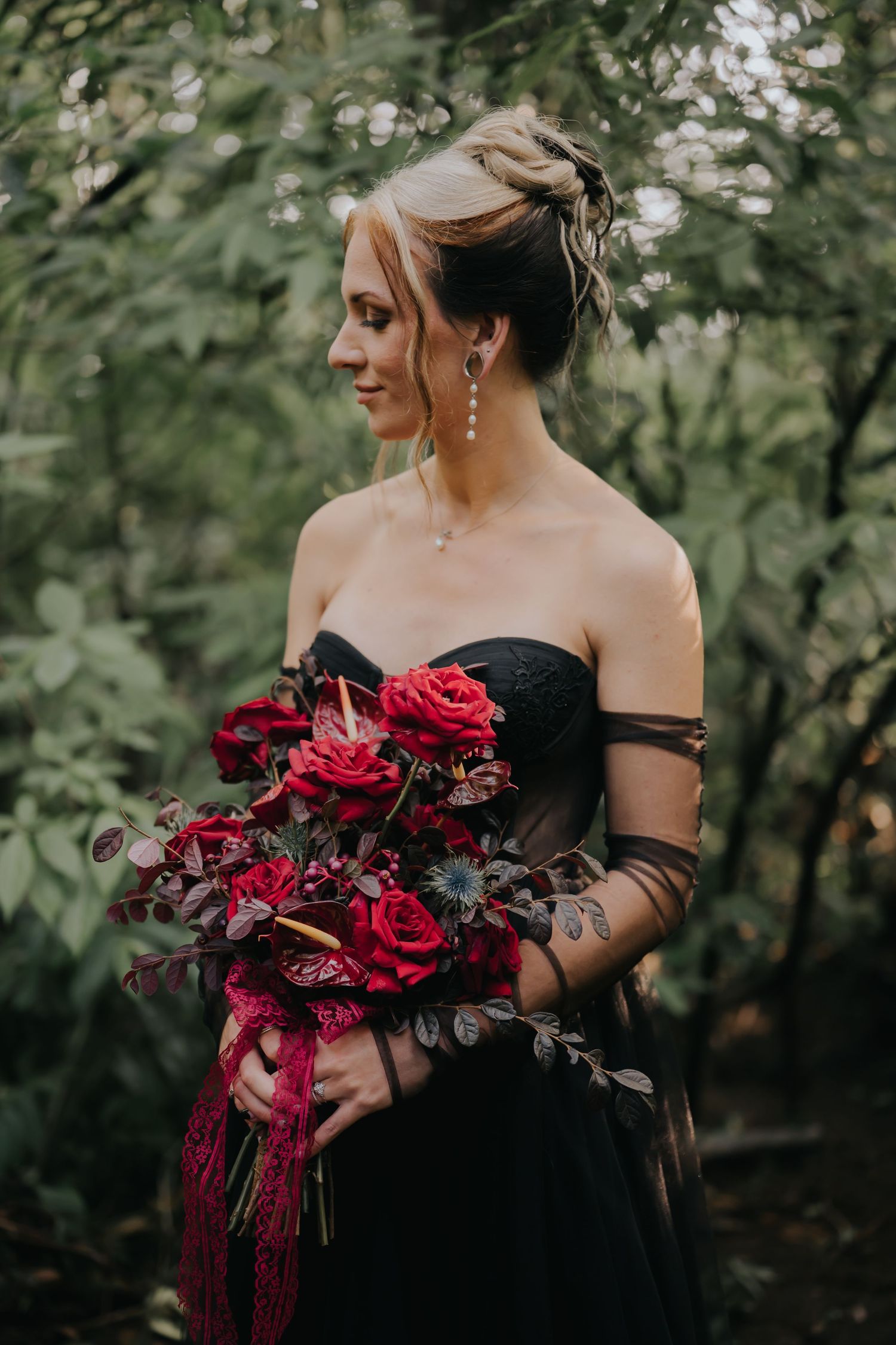 Person in black dress holding a dramatic bouquet of deep red roses against forest backdrop.