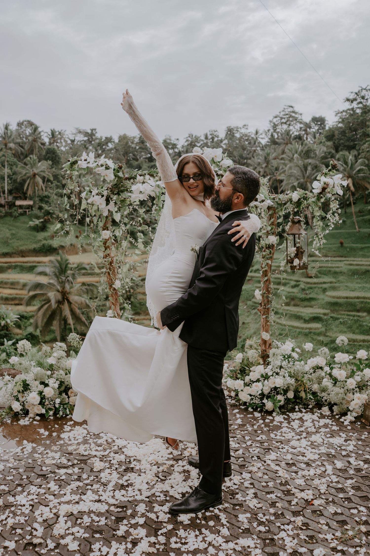A couple in wedding attire dances and spins together on a petal-covered ground in a garden setting.