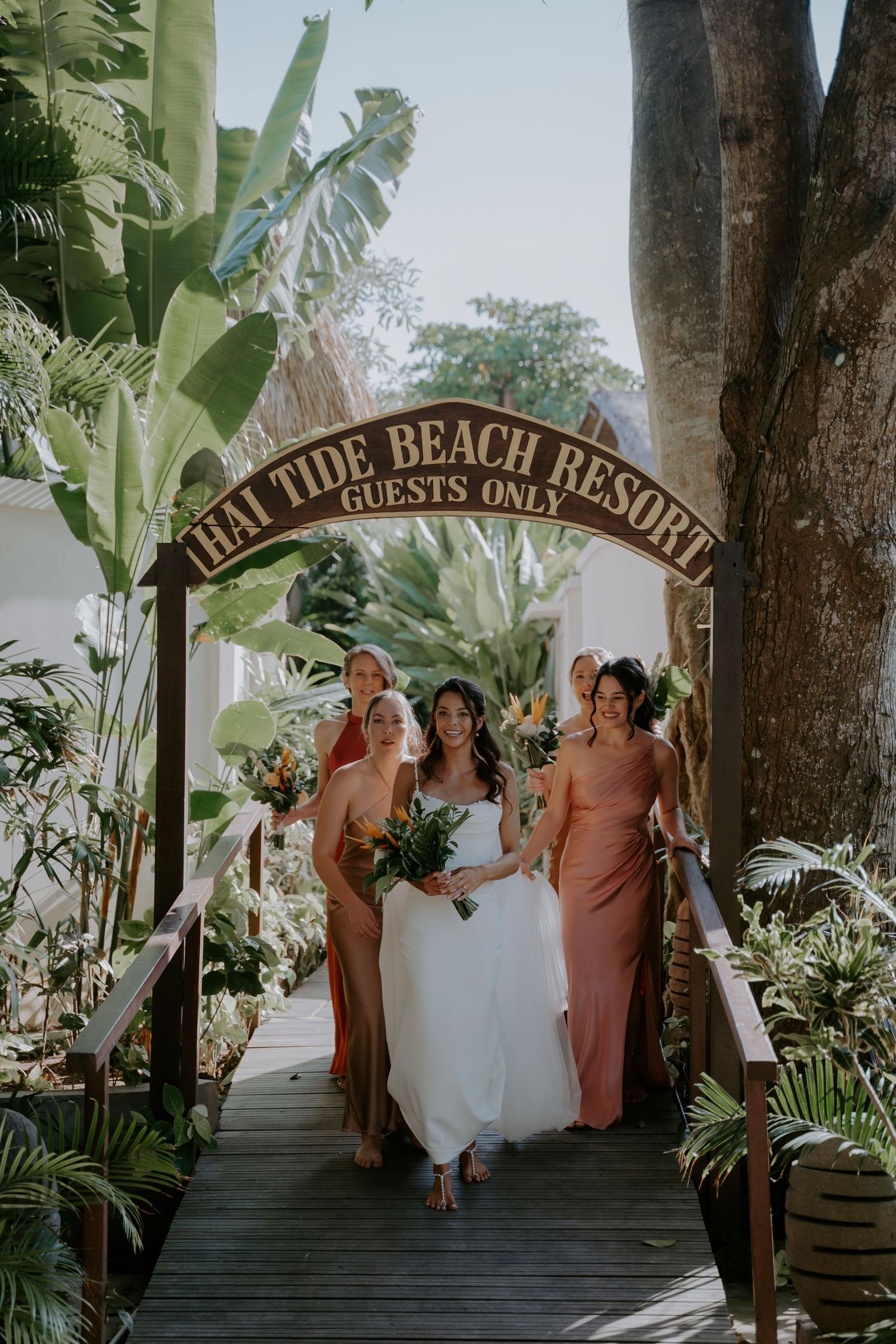 Wedding party poses under tropical palm trees and wooden arch at beachside resort entrance.