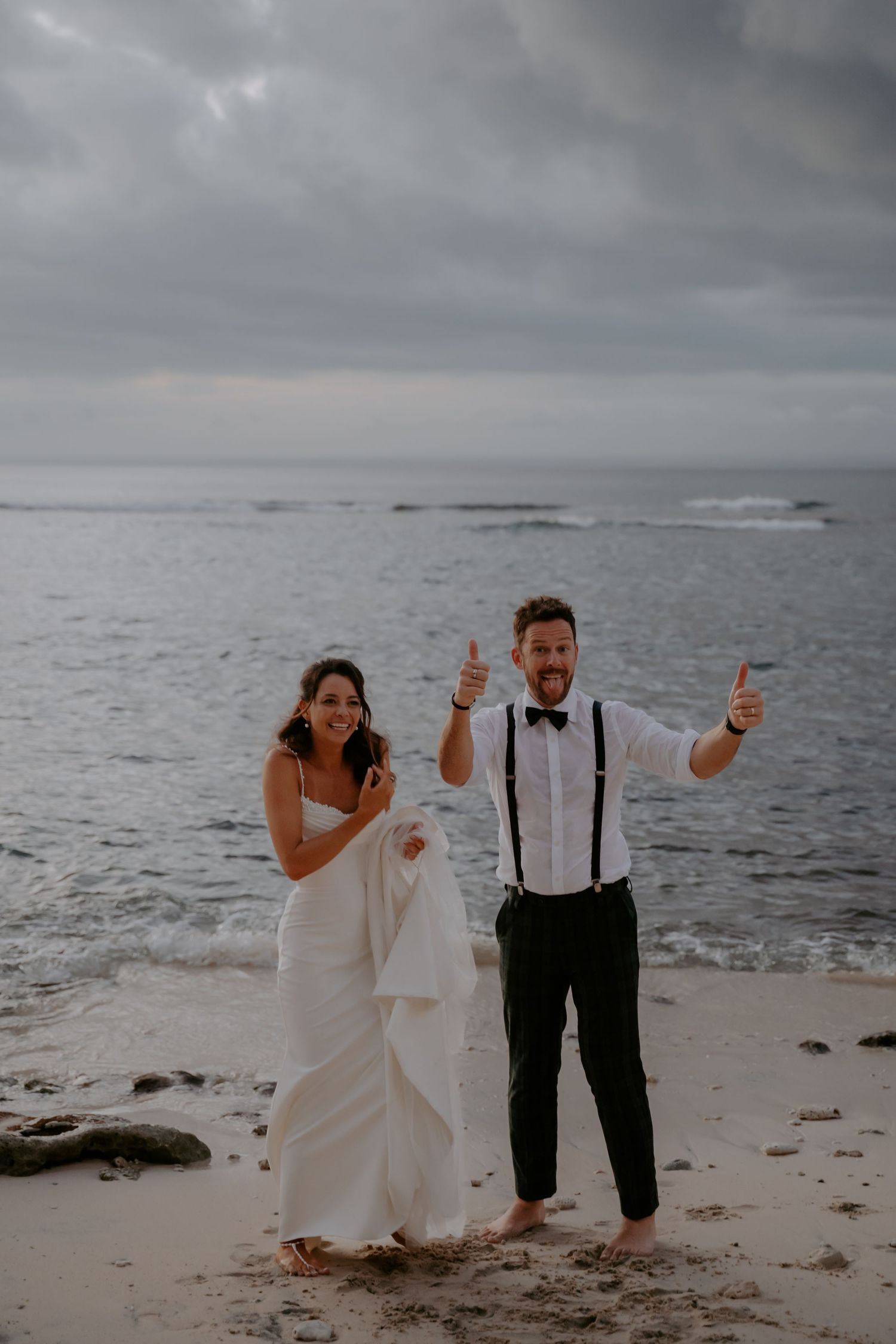 A couple poses playfully on a misty beach, celebrating their wedding day against a dramatic ocean backdrop.