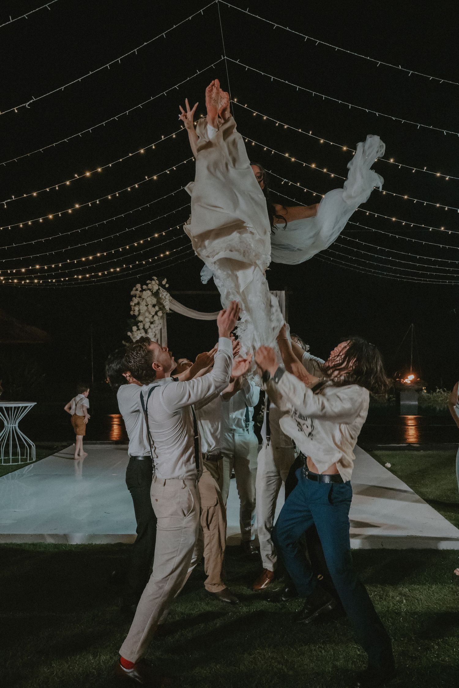 Wedding guests dance joyfully under twinkling string lights at an outdoor nighttime reception.
