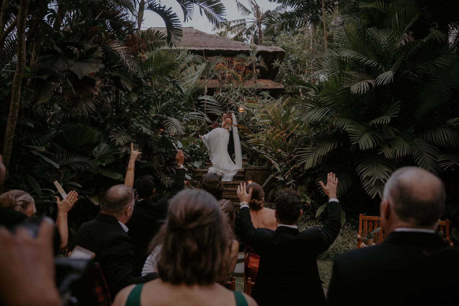 Wedding guests dance and celebrate at an outdoor reception venue with palm trees in the background.
