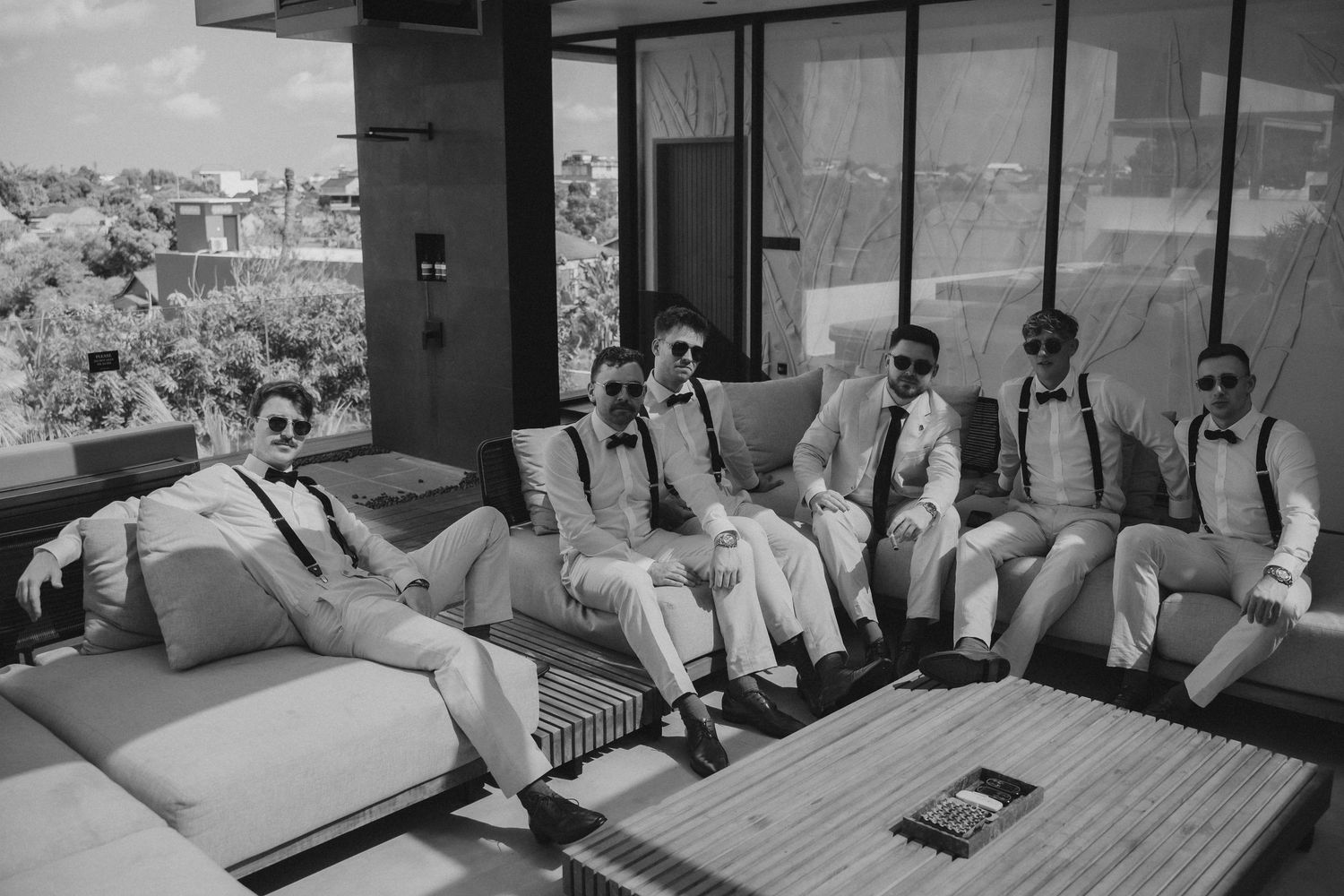 A wedding party relaxes on outdoor patio furniture in a black and white photograph.