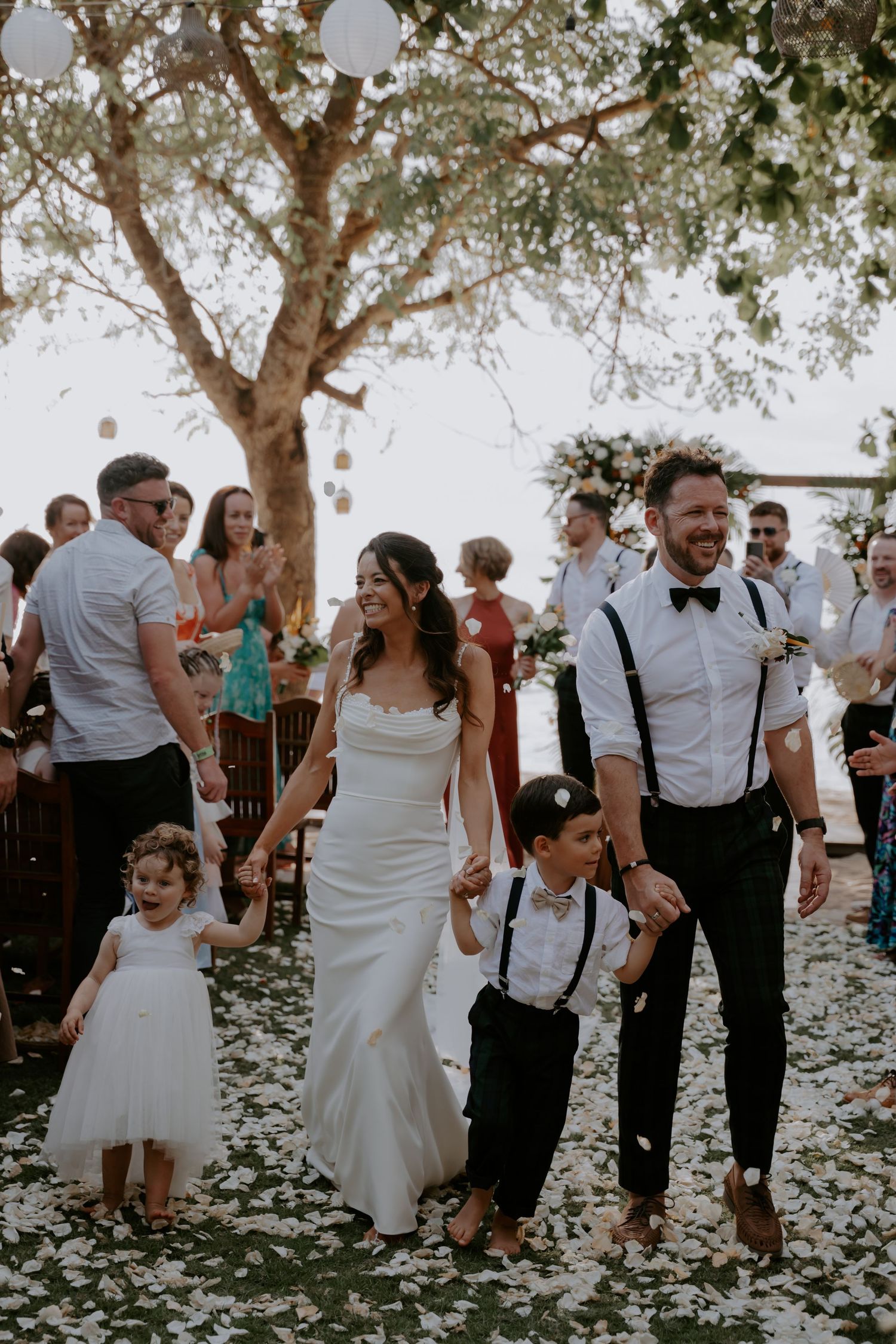 Wedding guests dance and celebrate under olive trees at an outdoor reception with white petals scattered on the ground.