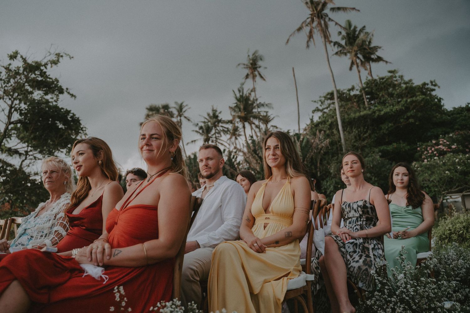 A group of people in colorful dresses and swimwear sit outdoors with palm trees in the background during sunset.