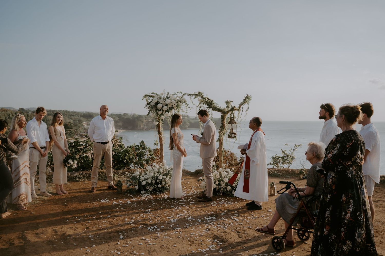 Wedding guests in white attire gather on a scenic coastal cliff overlooking the ocean during a sunset ceremony.