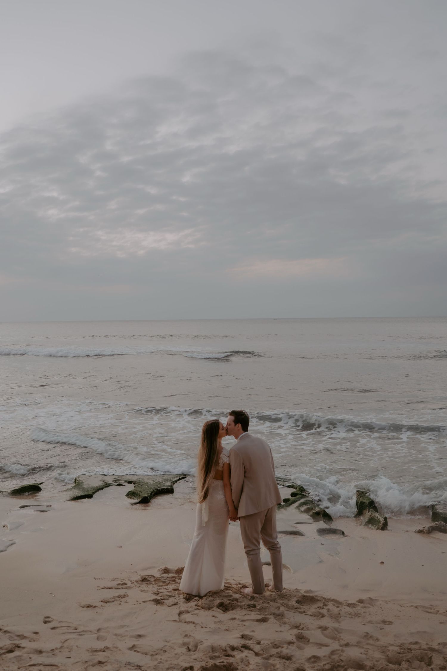 A couple walks along a misty beach at sunset with waves crashing on mossy rocks in the background.