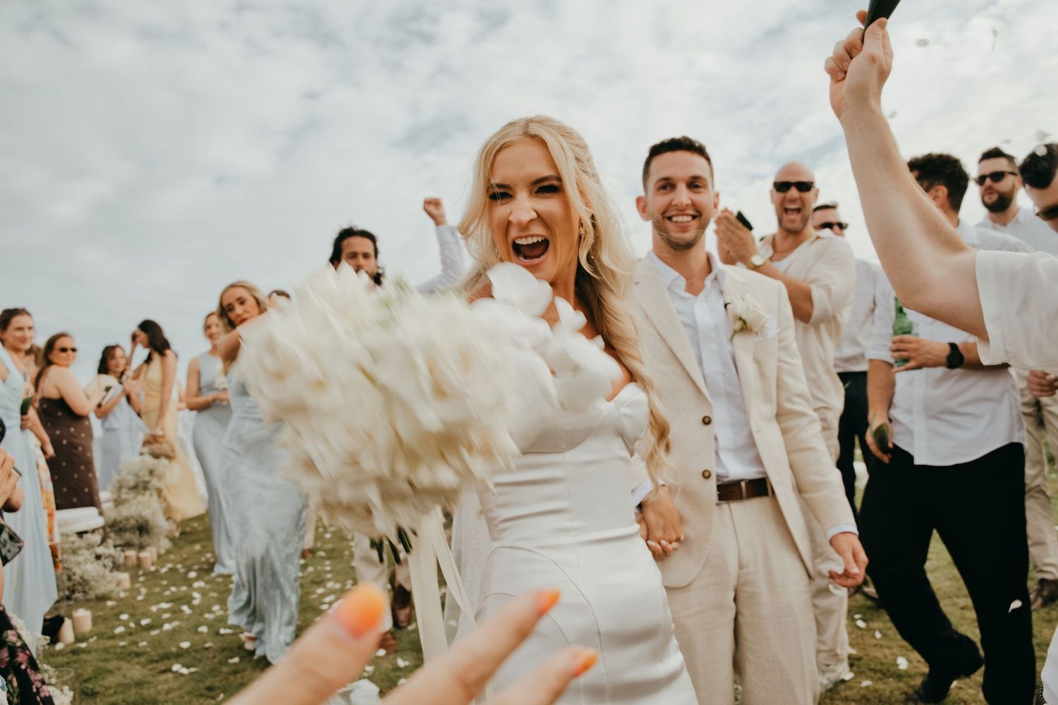 Joyful bride and groom celebrate outdoors with guests at a sunny wedding reception, bride holding white pampas grass.