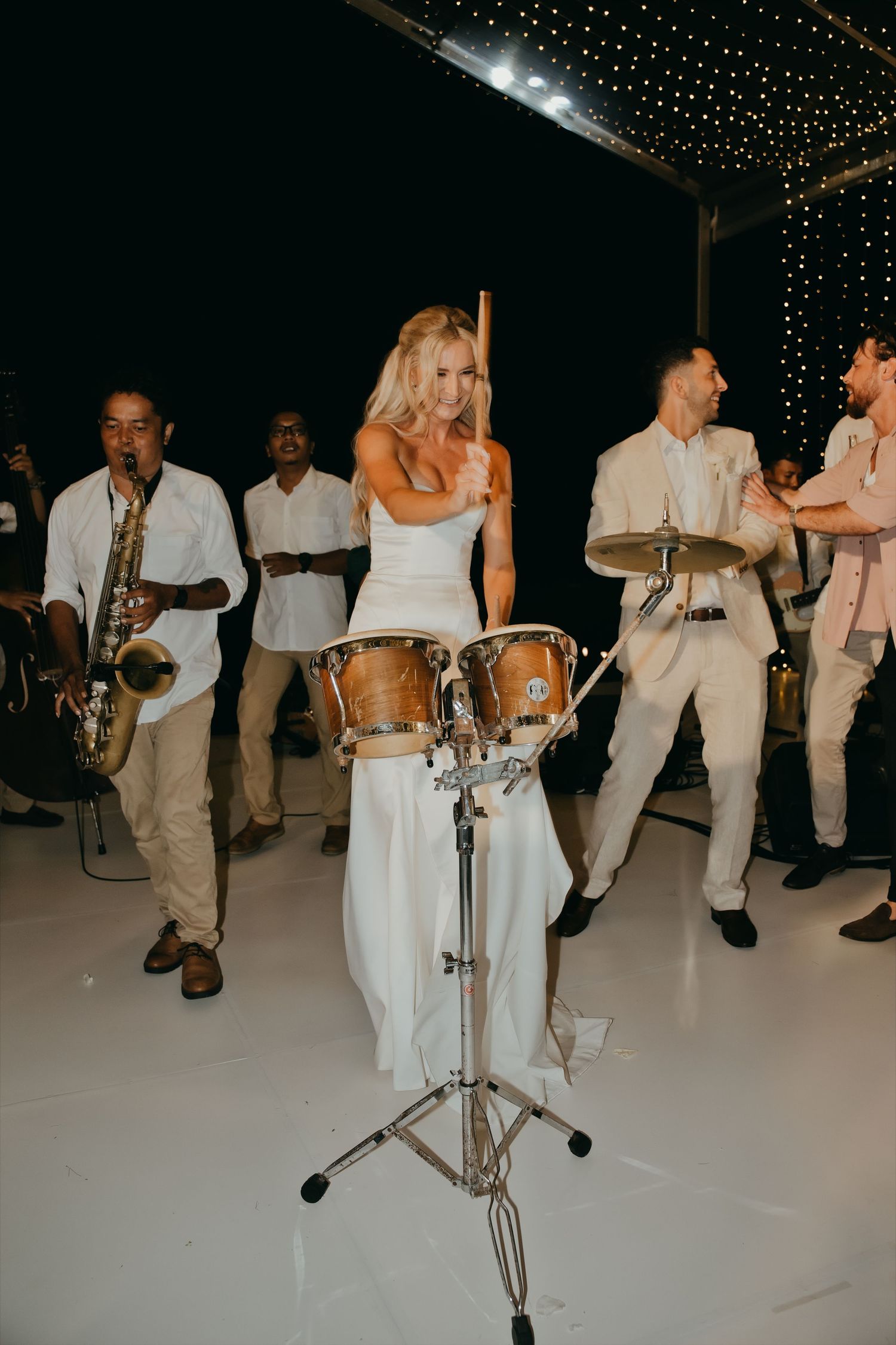 Bride in white gown plays bongo drums with live band at wedding reception celebration.