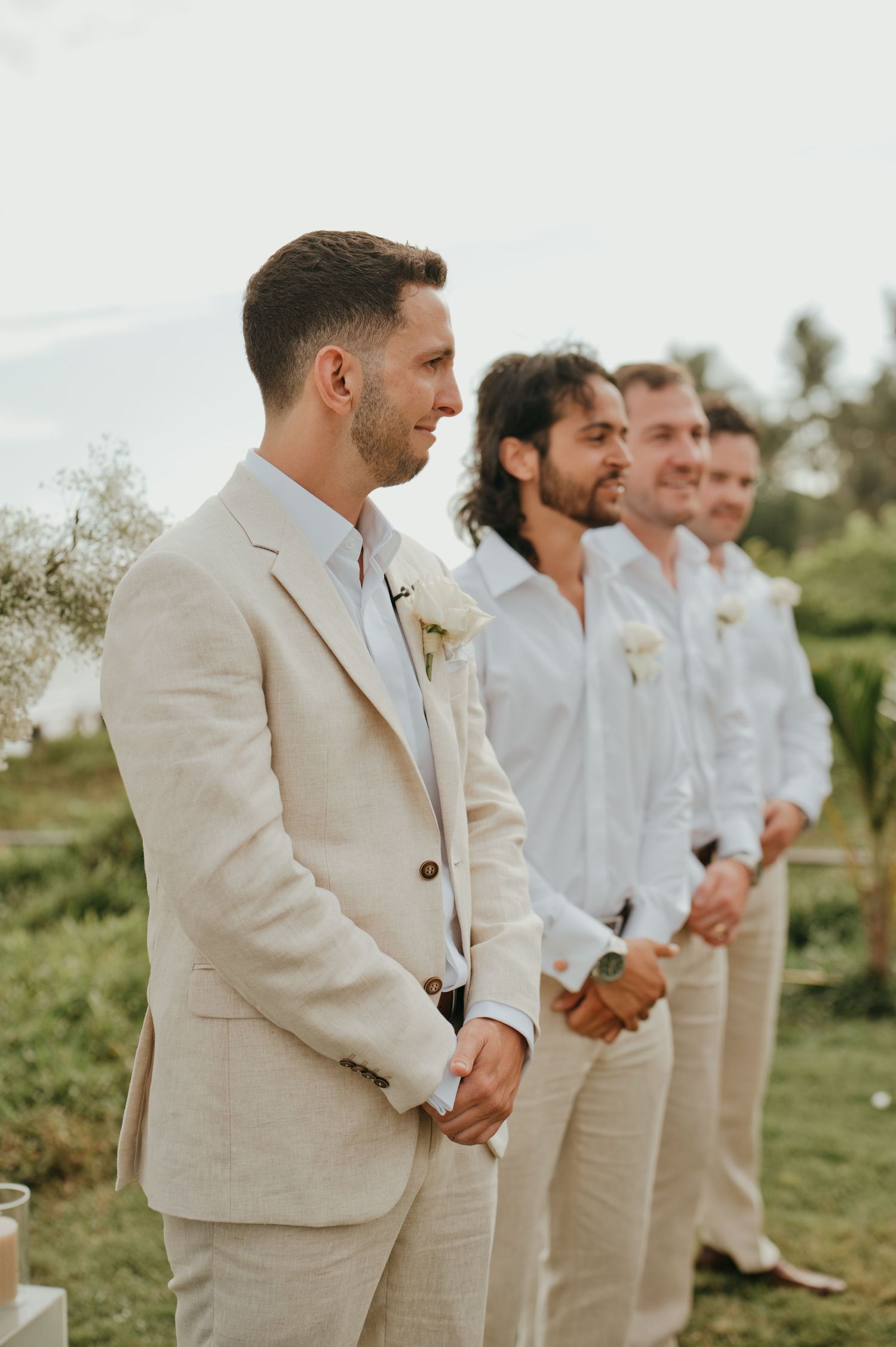 Groom in beige linen suit standing at the altar with groomsmen in white shirts at outdoor wedding.
