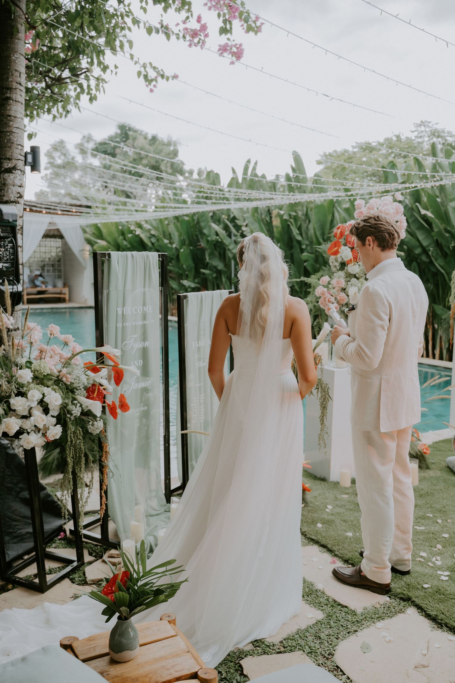 Bride and groom stand before a floral altar beside a pool with string lights overhead.