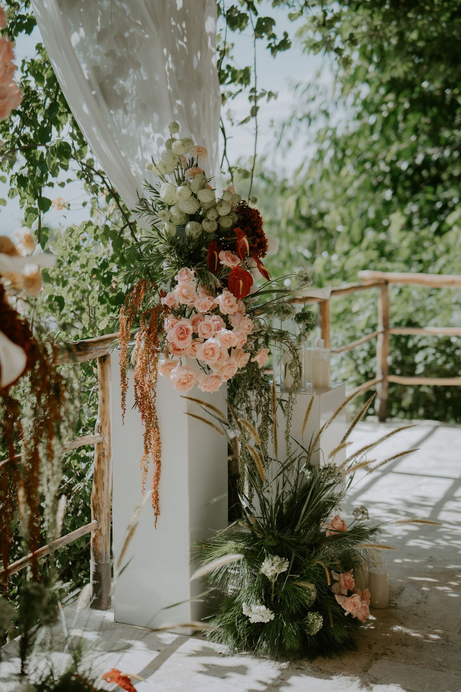 Wedding ceremony arch decorated with blush pink roses, burgundy flowers, and tropical greenery on outdoor terrace.