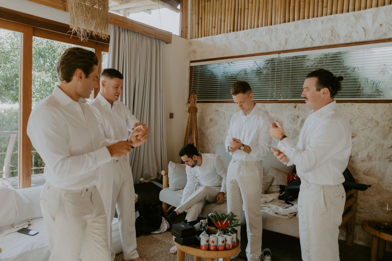 Groomsmen dressed in white attire chat and laugh together while getting ready in a tropical venue.