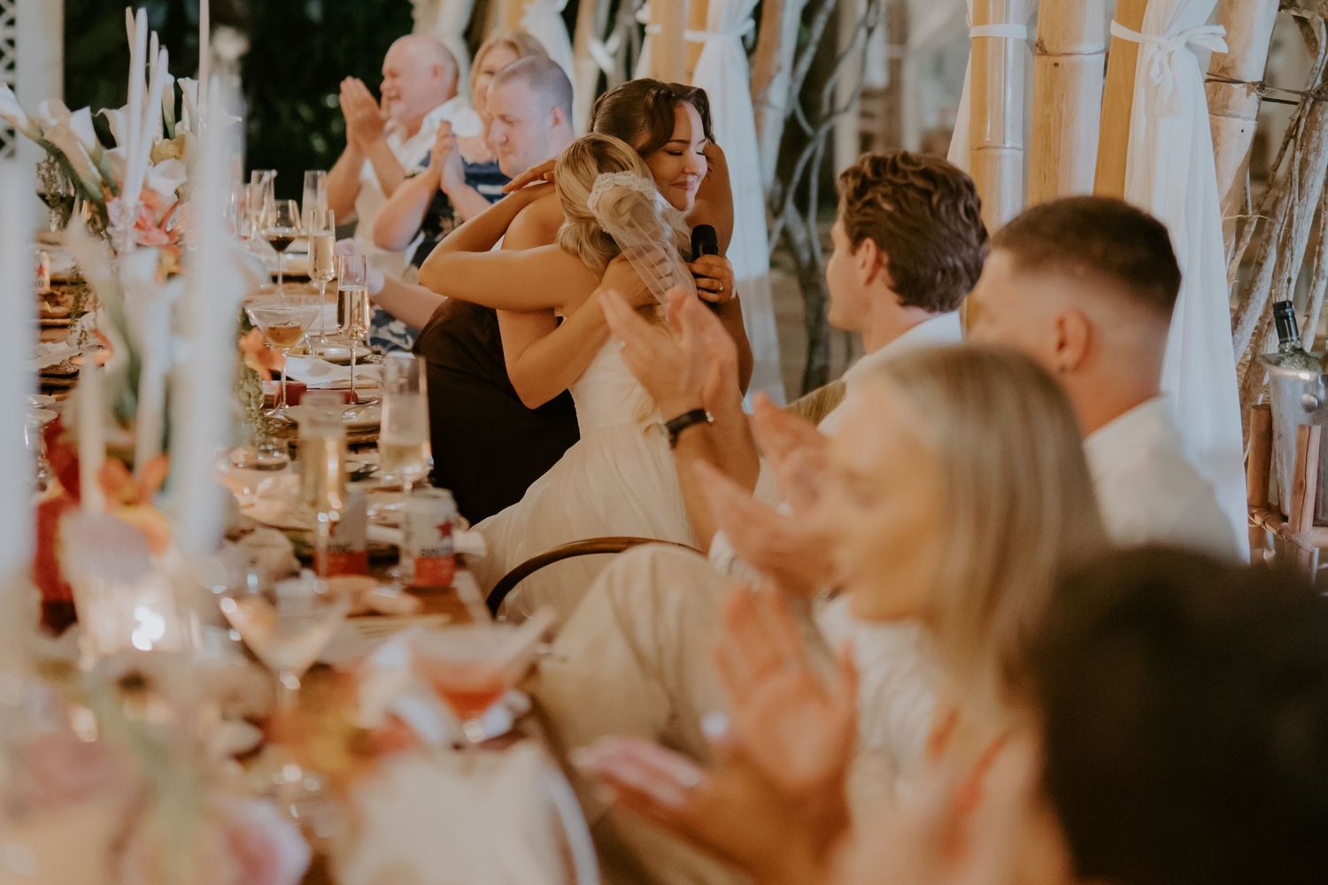 Bride embracing guest at wedding reception while guests applaud along decorated banquet table.