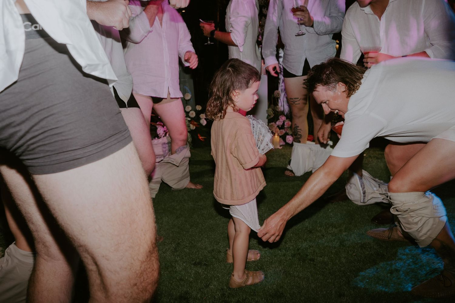 Young child dances with adult guest on grass at a lively outdoor wedding reception at night.