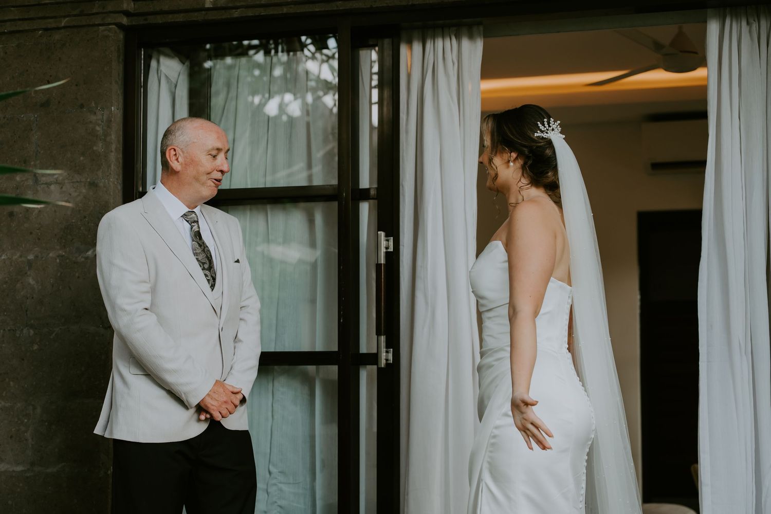 Bride in white gown and veil shares emotional first look moment with man in white suit at wedding venue.