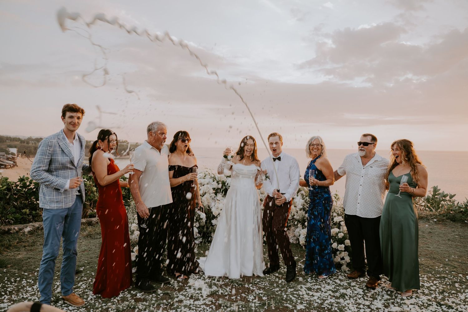 Wedding party celebrating with confetti streamers at a cliffside ceremony during a colorful sunset.
