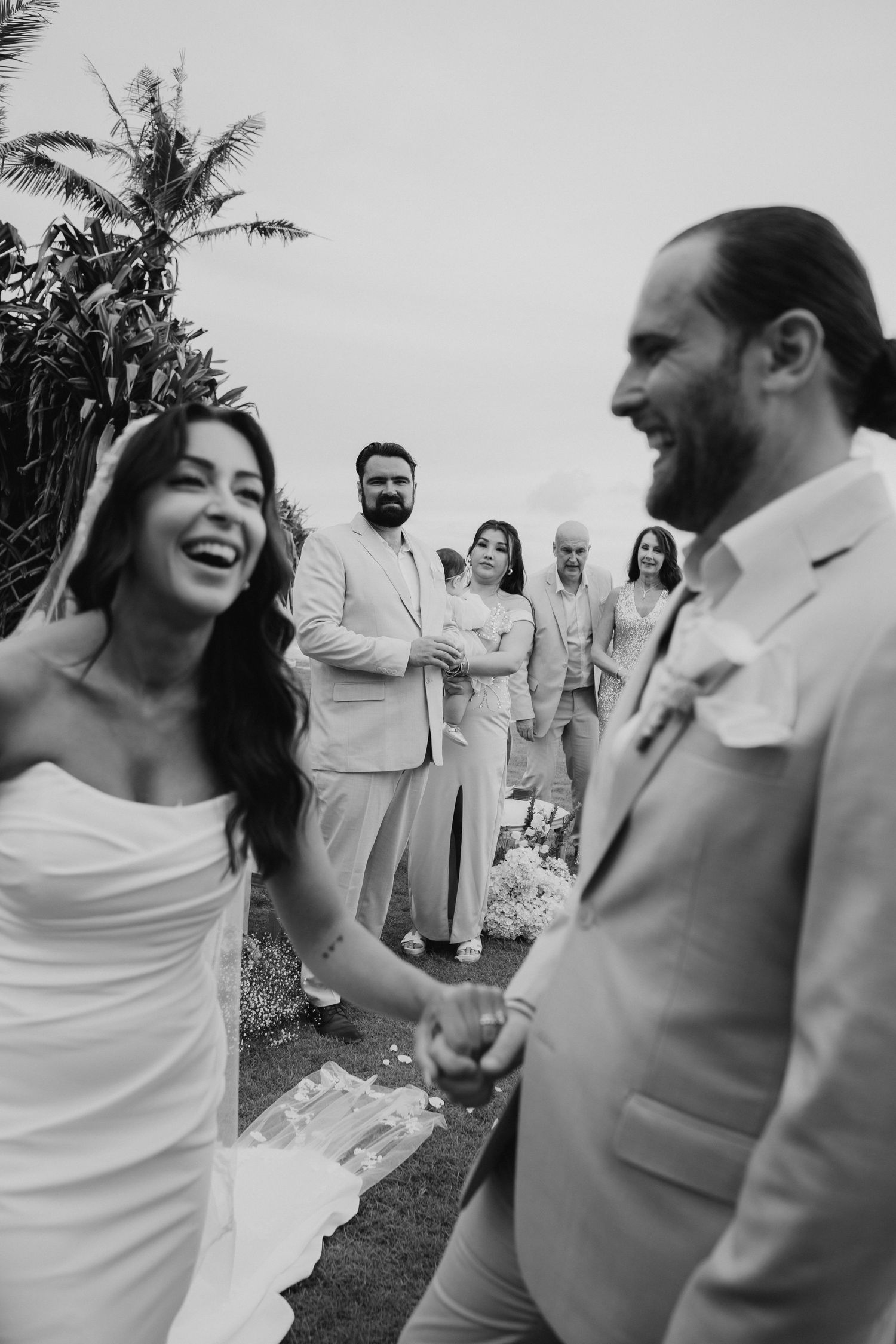 Black and white photo of laughing bride and groom walking past smiling wedding guests outdoors.