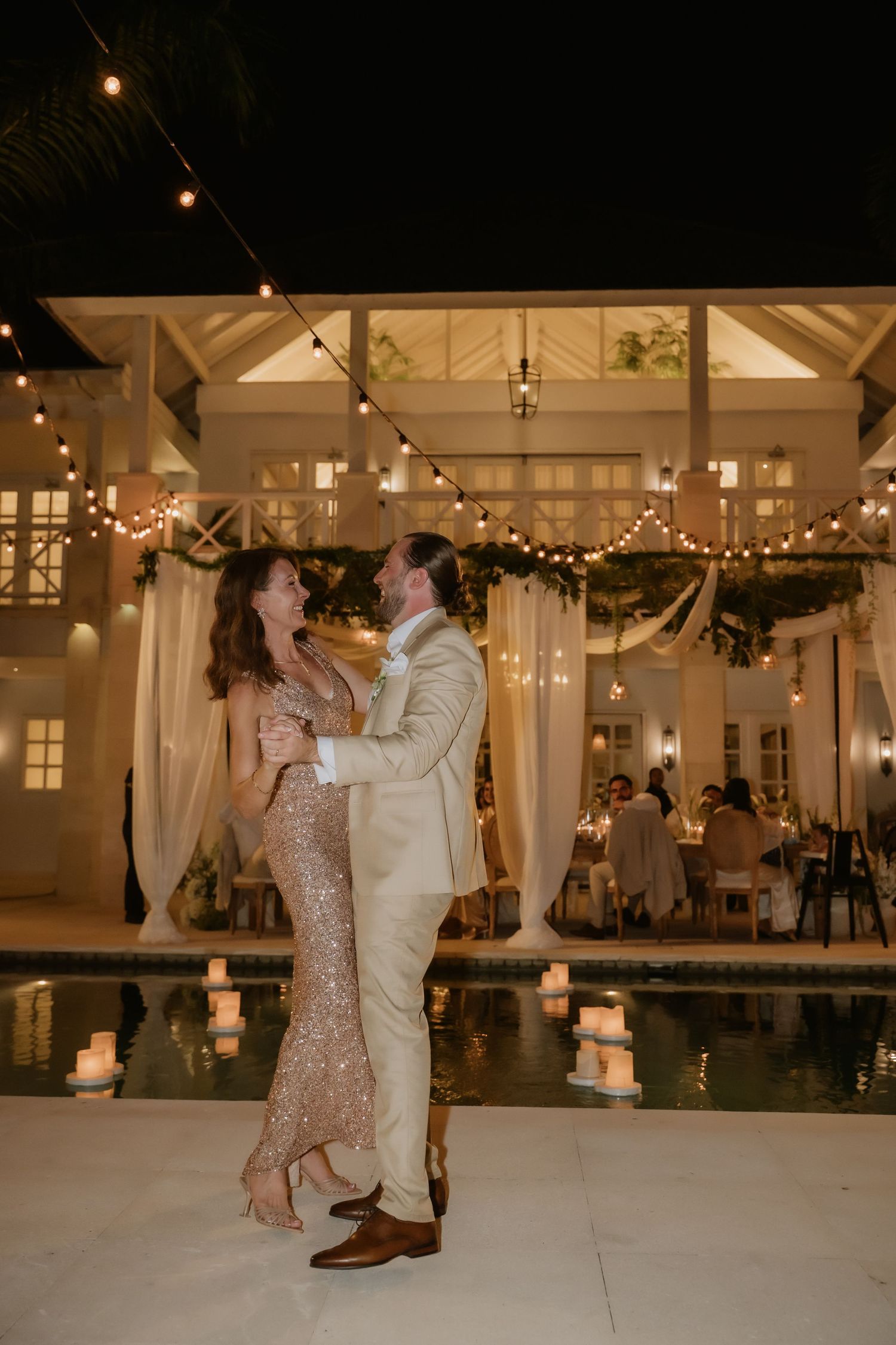 Bride and groom share first dance in sequin gown by candlelit pool under string lights at romantic outdoor wedding reception.