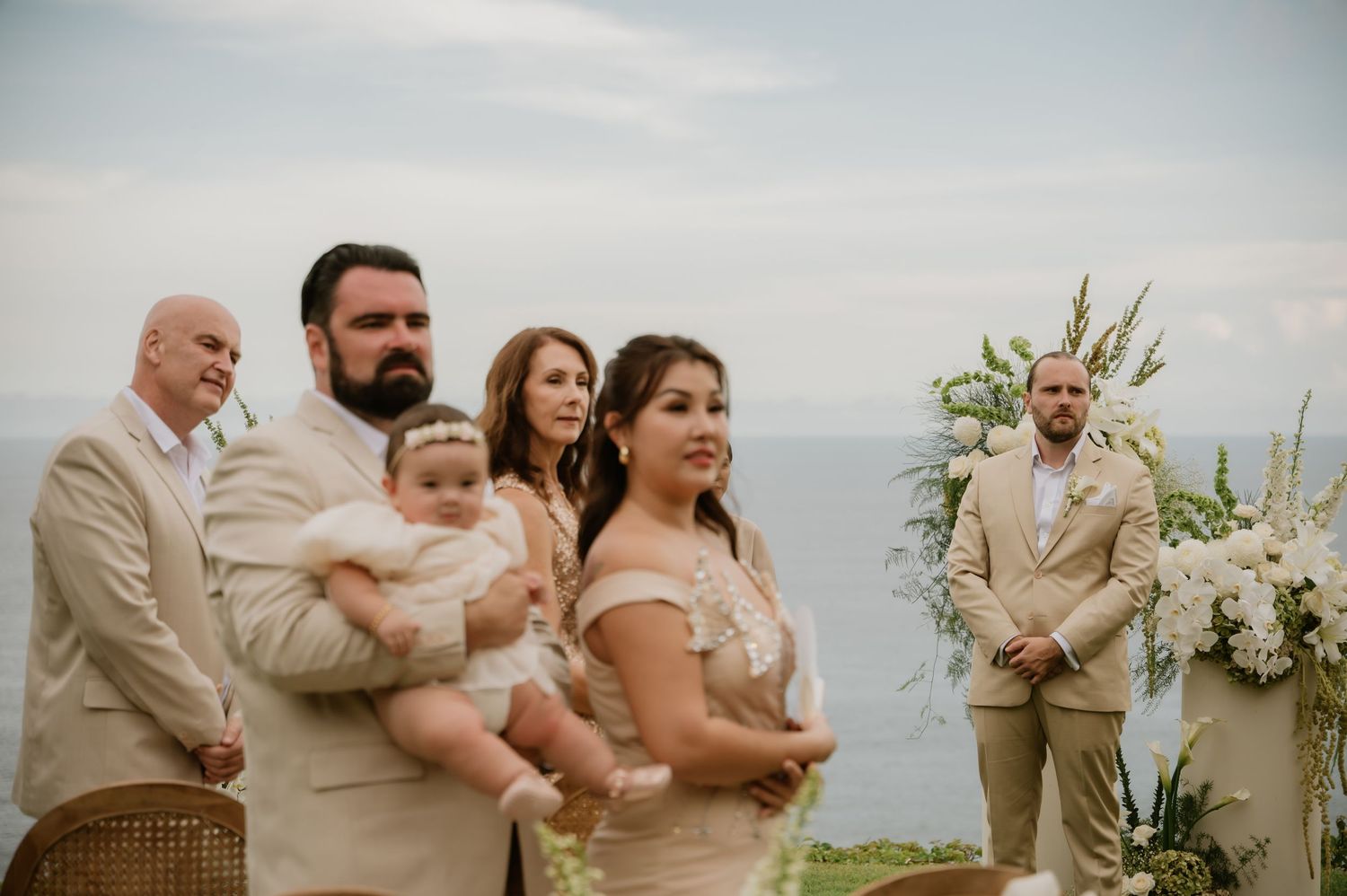 Family poses at outdoor wedding ceremony with ocean backdrop, dressed in elegant beige attire, holding a baby in white.