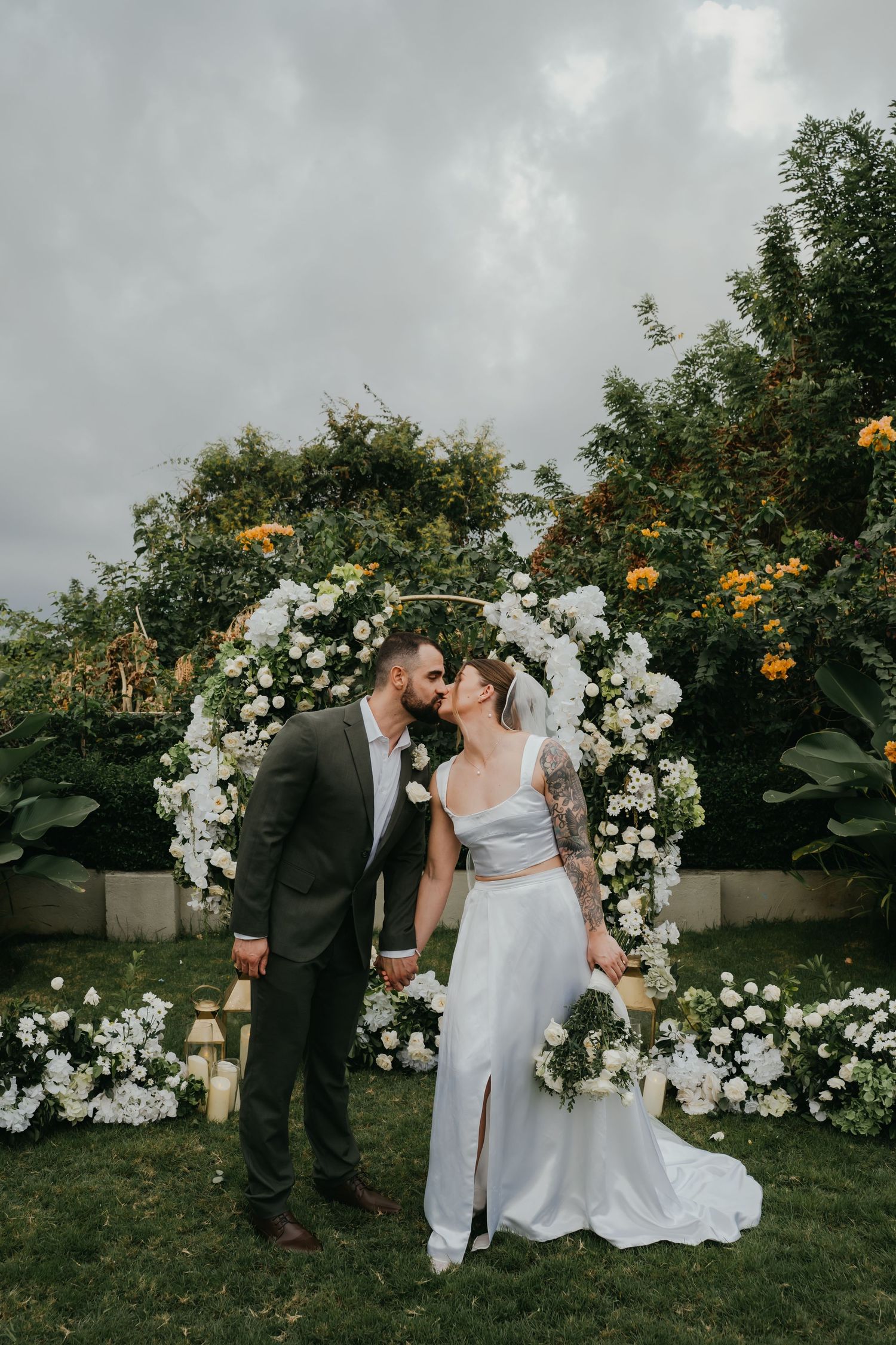 Newlyweds kiss in front of circular white floral arch at outdoor garden wedding ceremony.