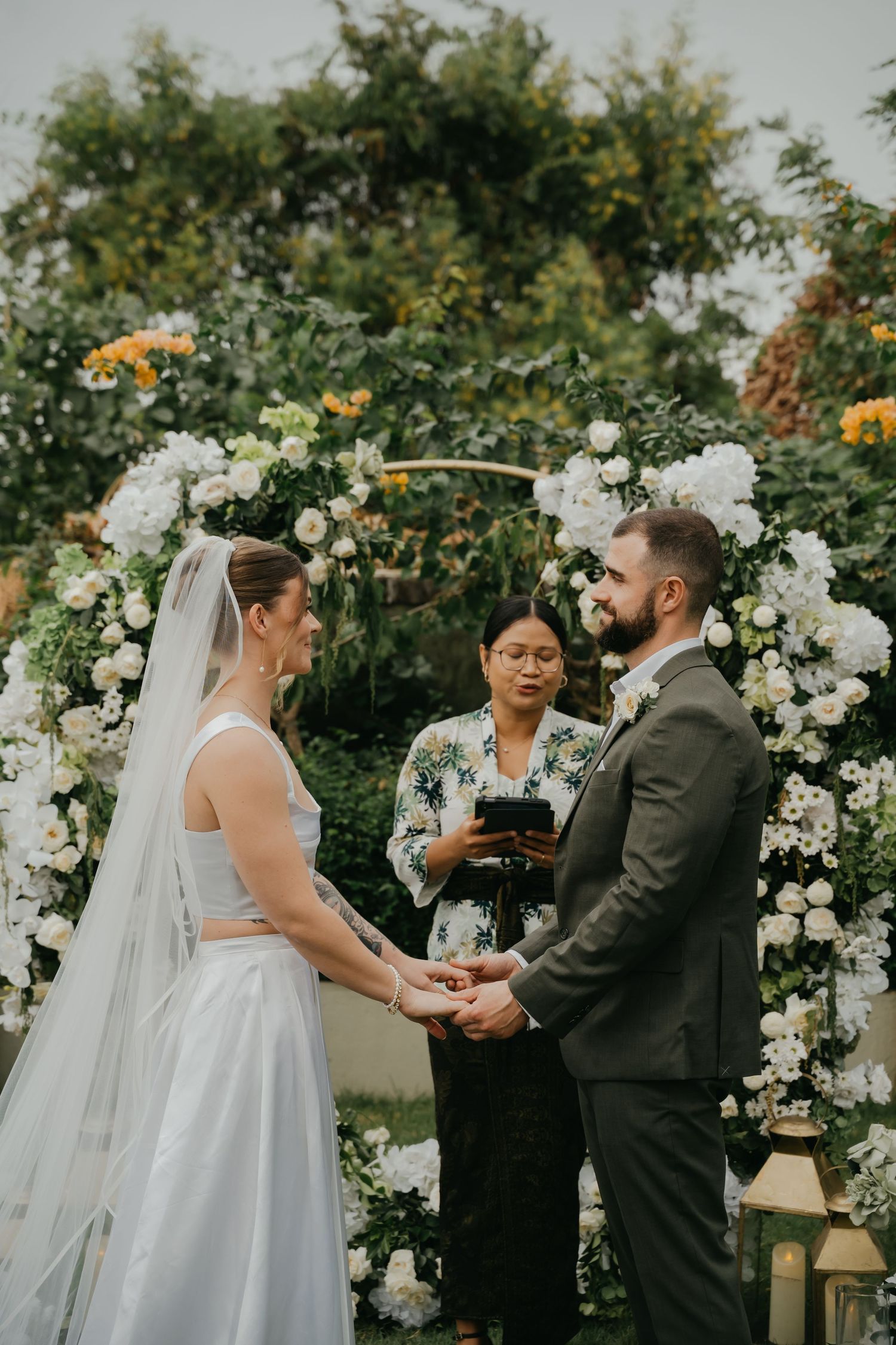 Bride and groom exchange vows under floral arch with white roses and greenery at outdoor wedding ceremony.