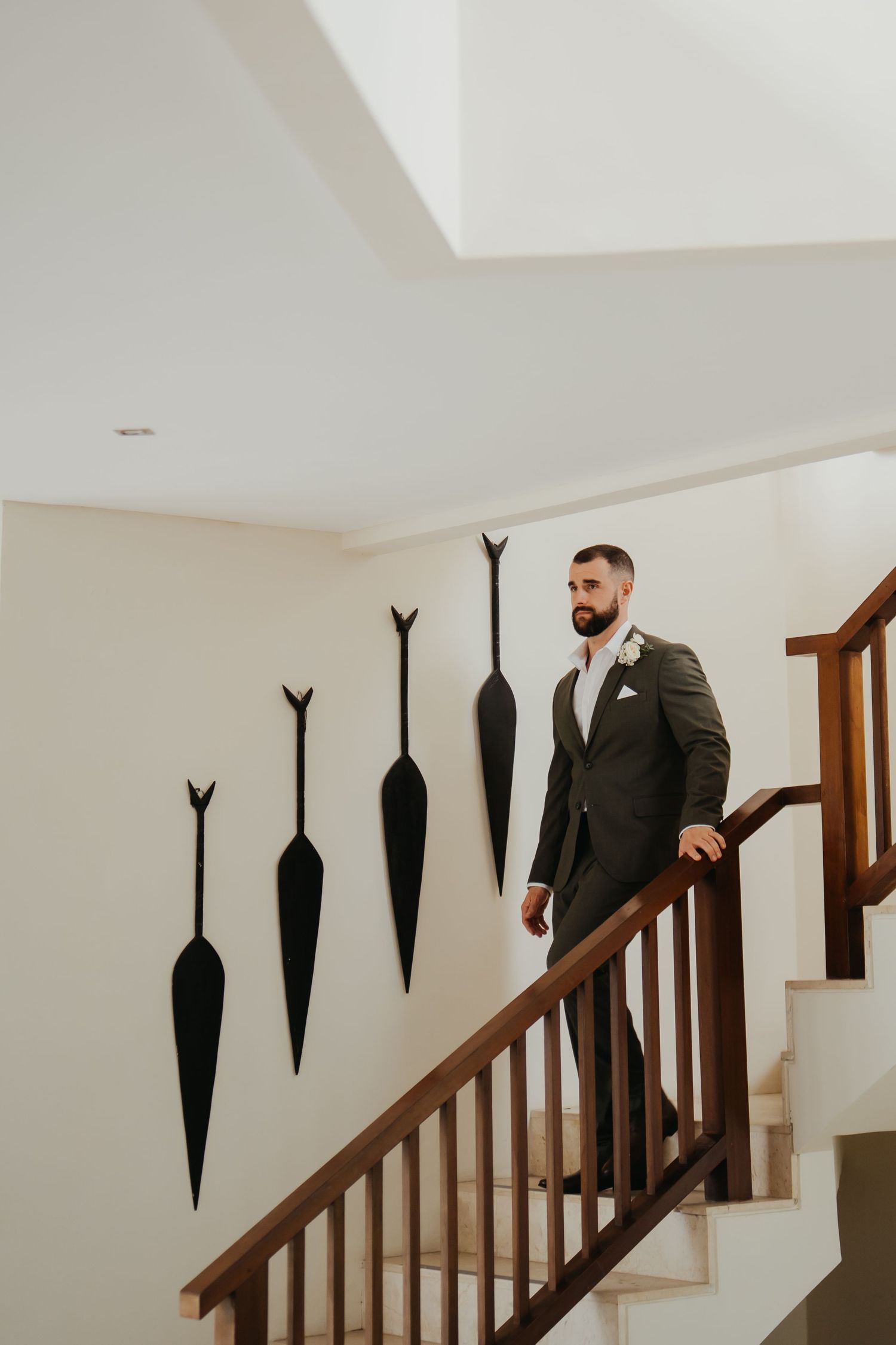 A dapper groom in a dark suit stands on wooden stairs beside decorative wall-mounted oar art.