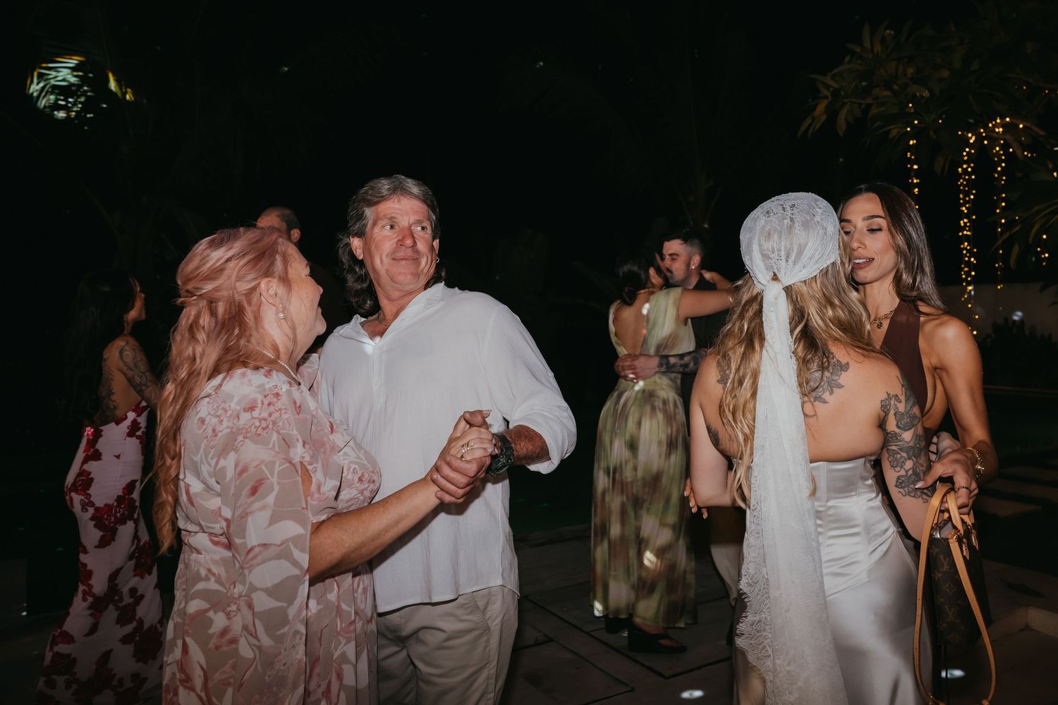 Guests dance joyfully at an outdoor evening wedding reception with string lights in the background.