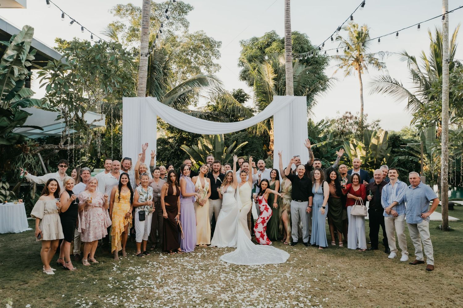 Wedding party and guests celebrate with confetti under a white floral arch at a tropical outdoor venue.