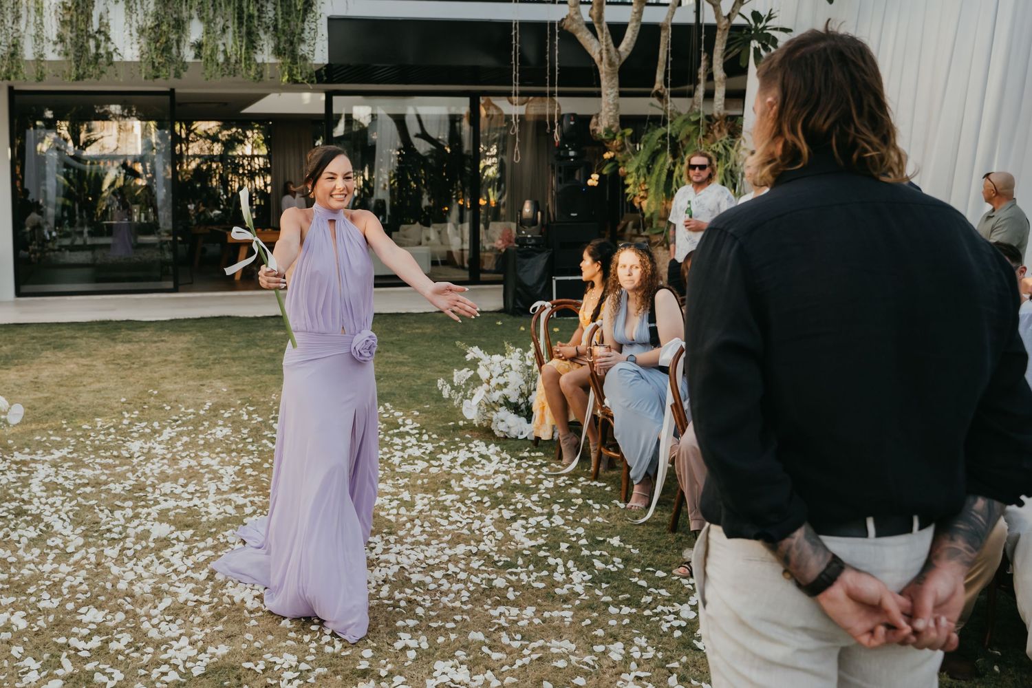 Bridesmaid in lavender gown tossing flower petals at outdoor wedding ceremony.