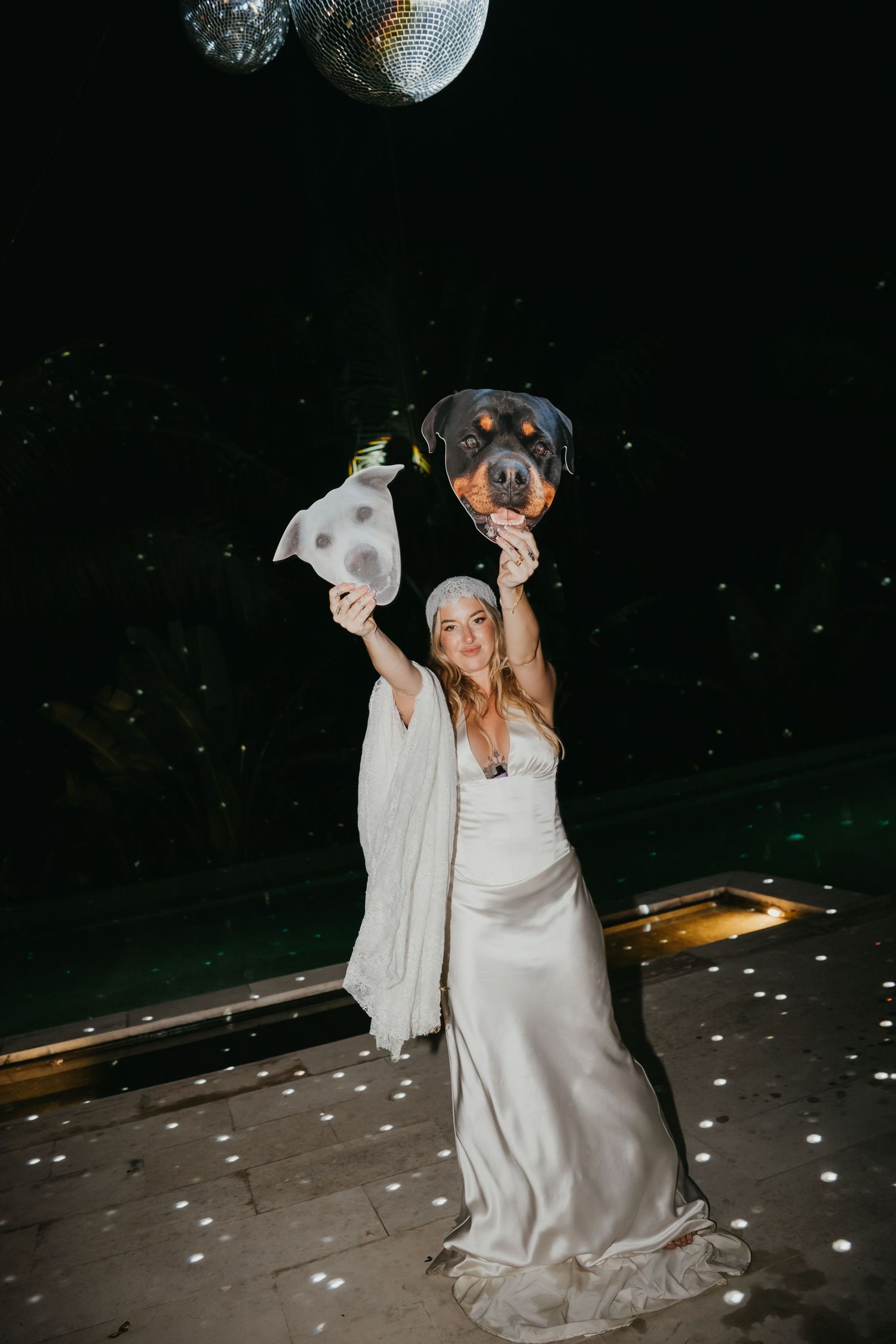 Bride in white satin gown holds up two dog face cutouts under a disco ball at her wedding reception.