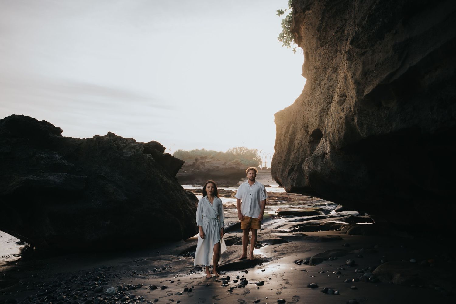 A couple stands silhouetted in a dramatic rocky sea cave with sunlight streaming through the opening.