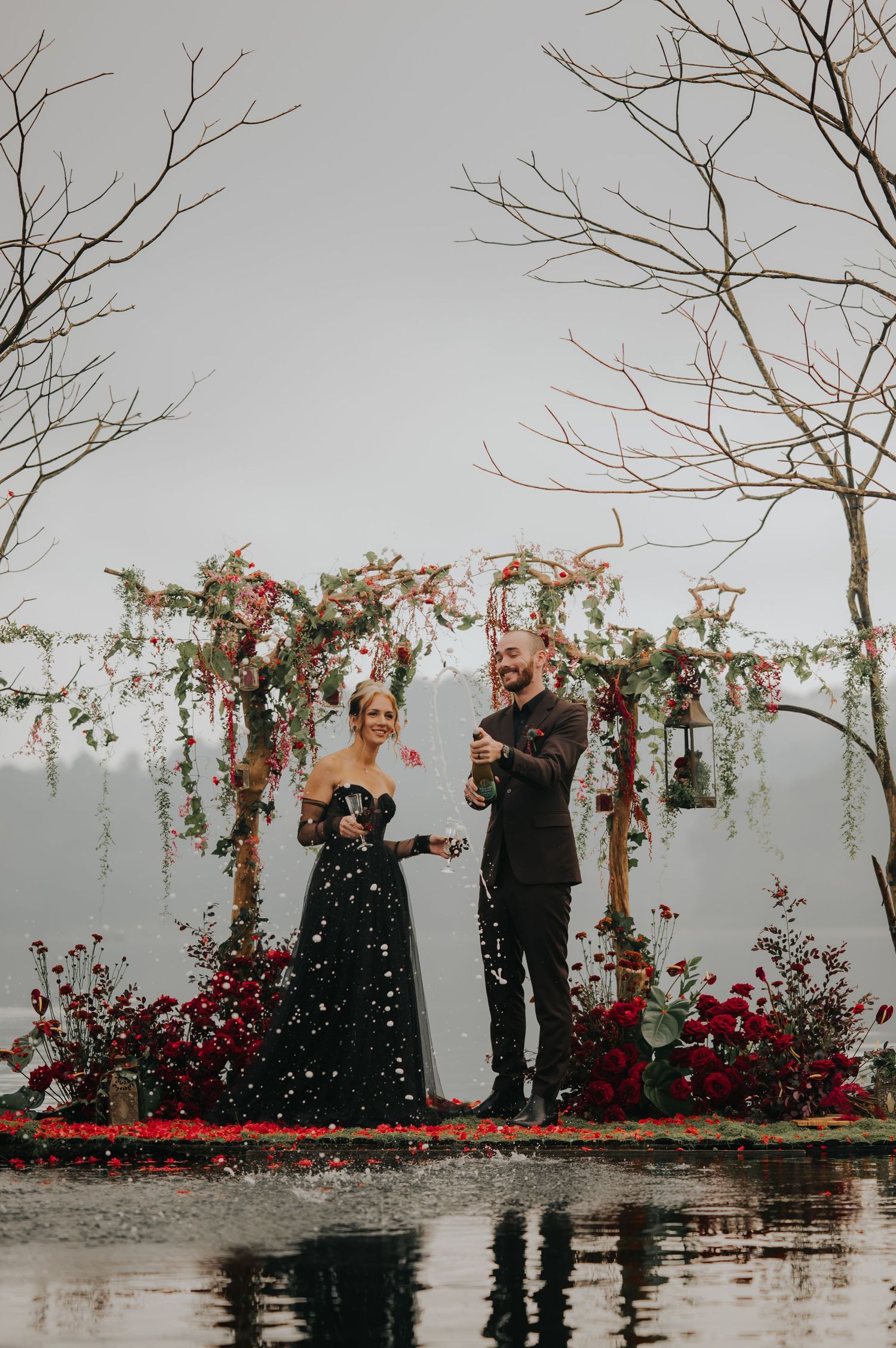 Couple celebrating on a water platform decorated with red flowers and bare branches against foggy backdrop.