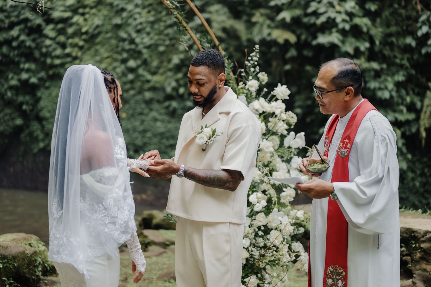 Groom places ring on brides finger during outdoor wedding ceremony officiated by priest in red stole.