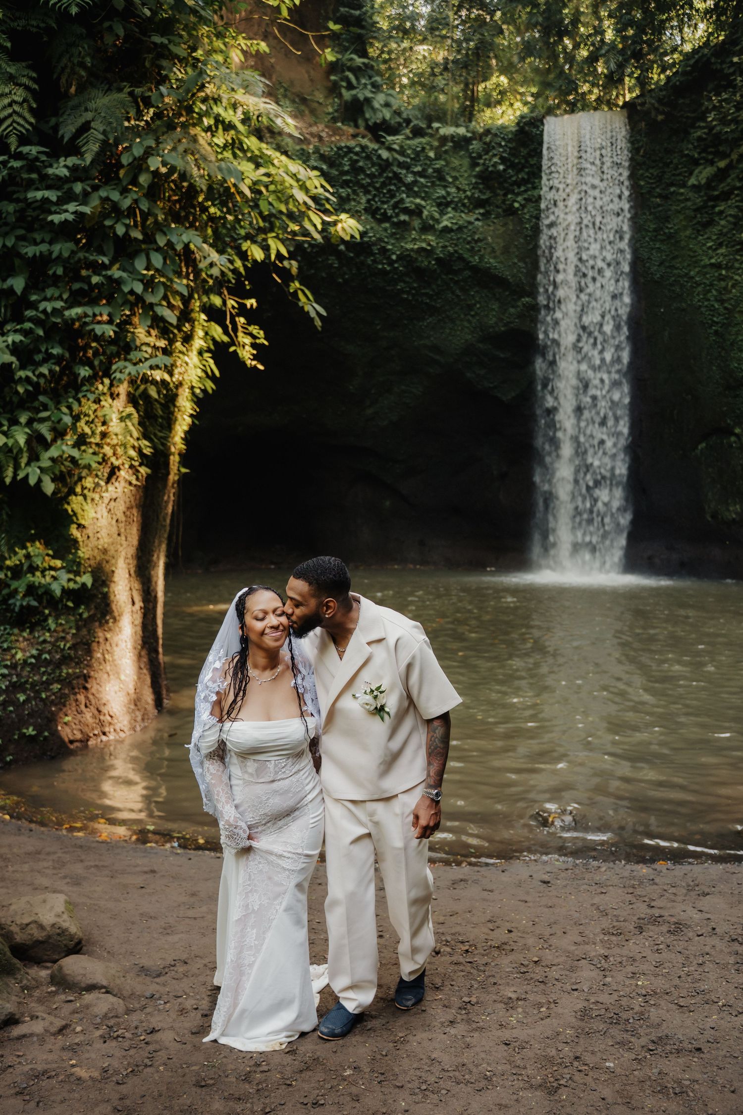 Newlywed couple embraces lovingly in front of a serene waterfall surrounded by lush green jungle foliage.